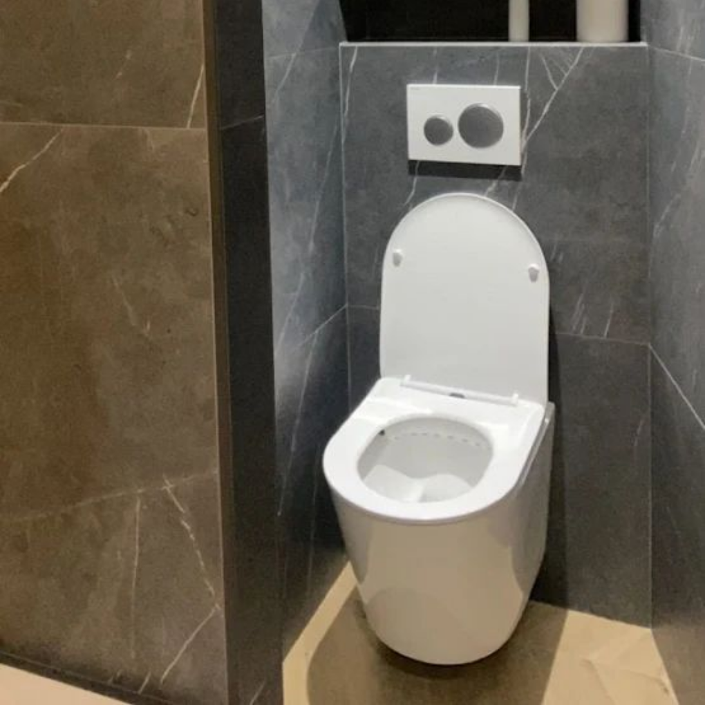 Toilettes blanches modernes dans une cabine de salle de bains carrelée de gris, avec un bouton de double chasse d'eau au-dessus.