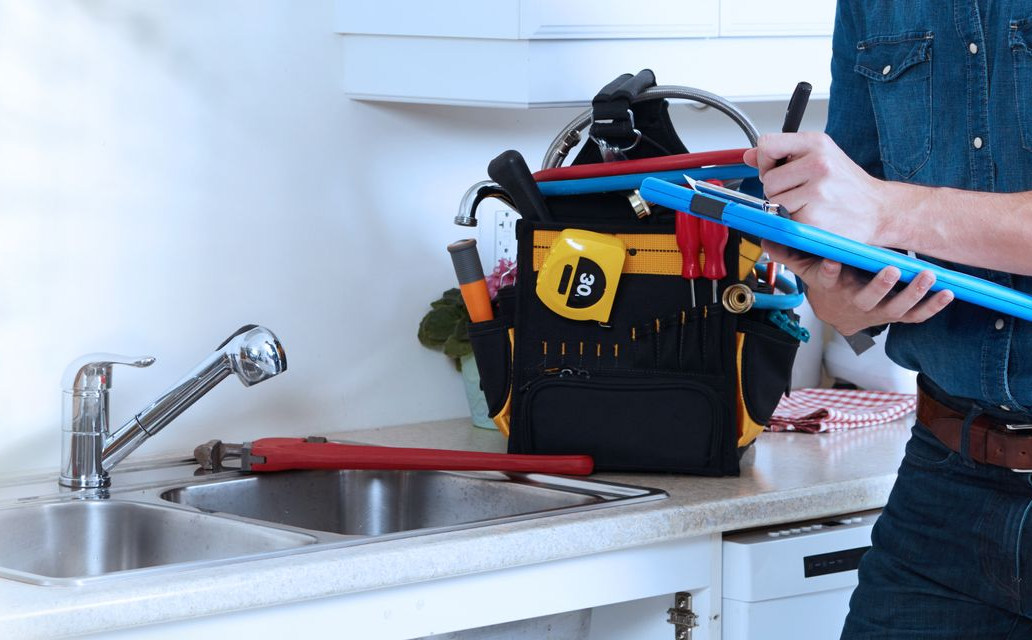 Une personne portant une boîte à outils et un niveau bleu se tient à côté d'un évier de cuisine, avec un tuyau rouge sur le comptoir.