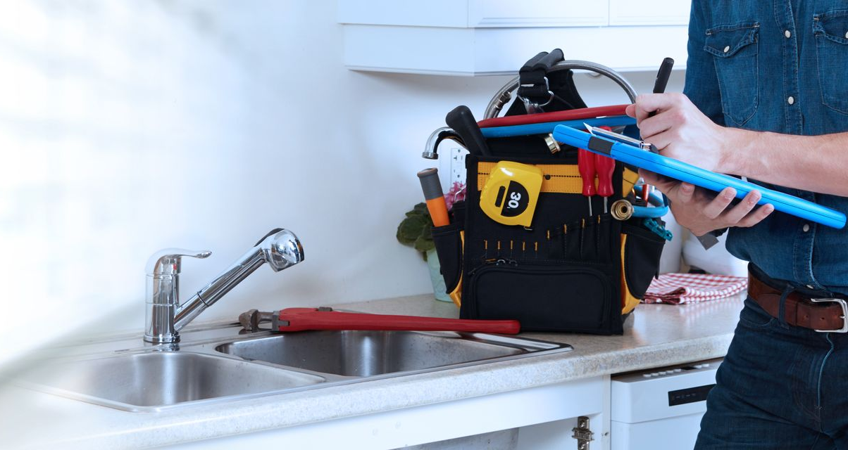 Un plombier répare un évier à l'aide d'une clé, tandis qu'une trousse à outils et un mètre ruban sont posés sur le comptoir.