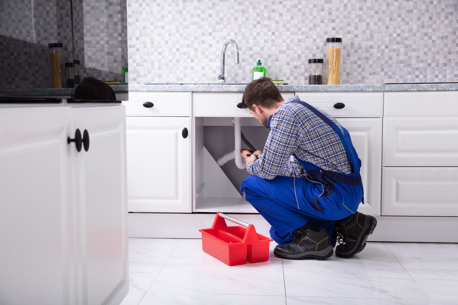 Une personne agenouillée dans une cuisine, réparant un lave-vaisselle ouvert, une boîte à outils rouge à proximité.