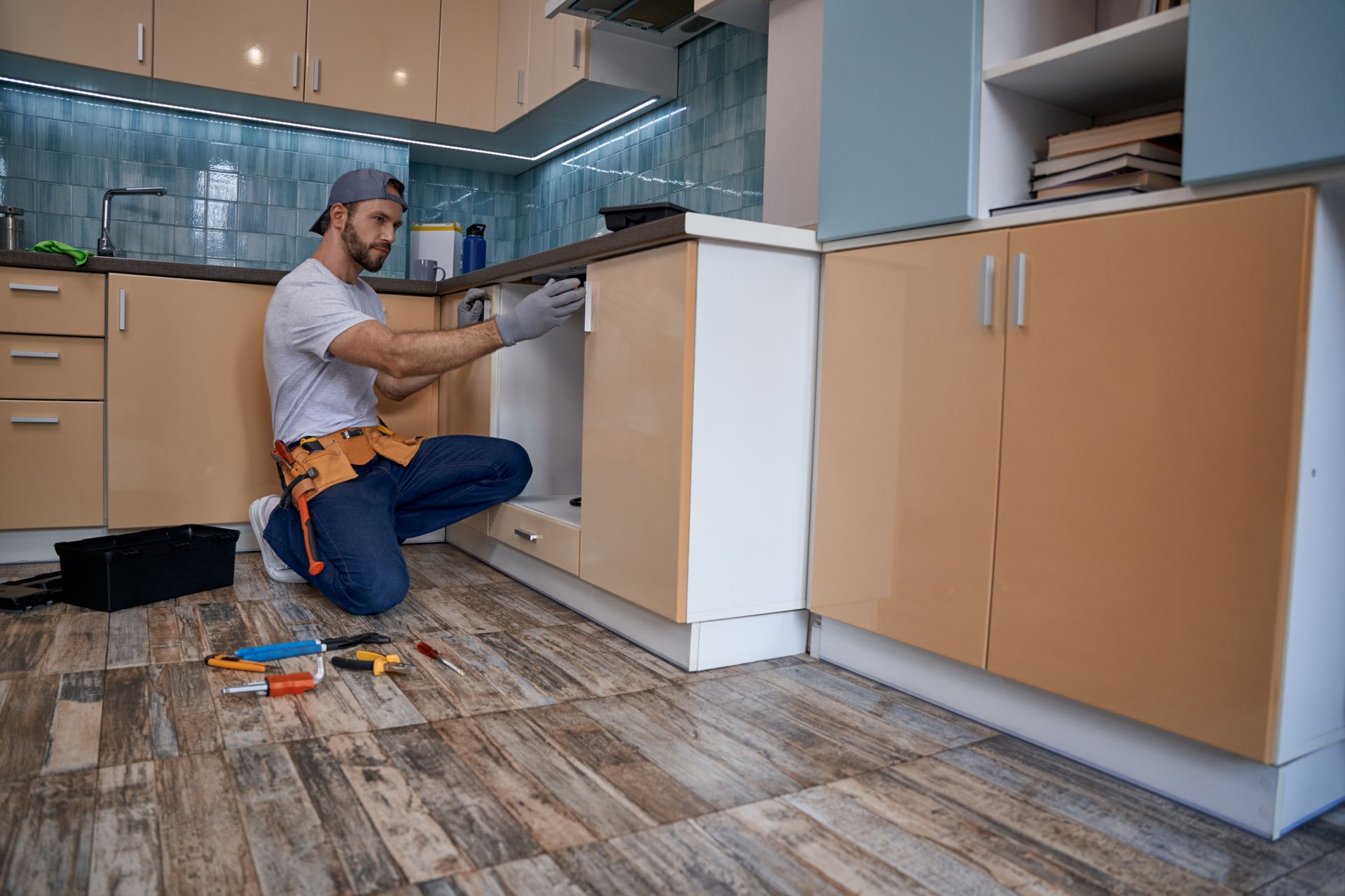 Un ouvrier installe des armoires de cuisine dans une cuisine moderne, agenouillé avec ses outils sur le sol.