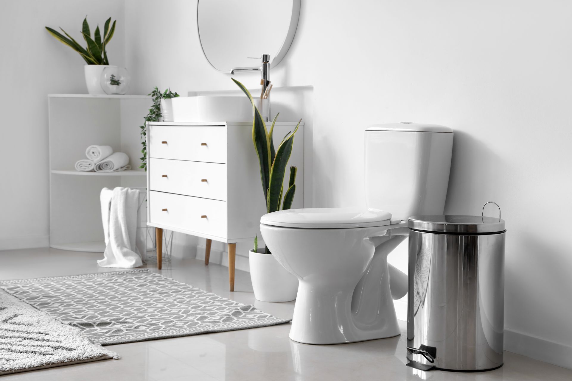 Salle de bains moderne blanche avec toilettes, lavabo, baignoire, poubelle chromée et plantes sur un tapis gris