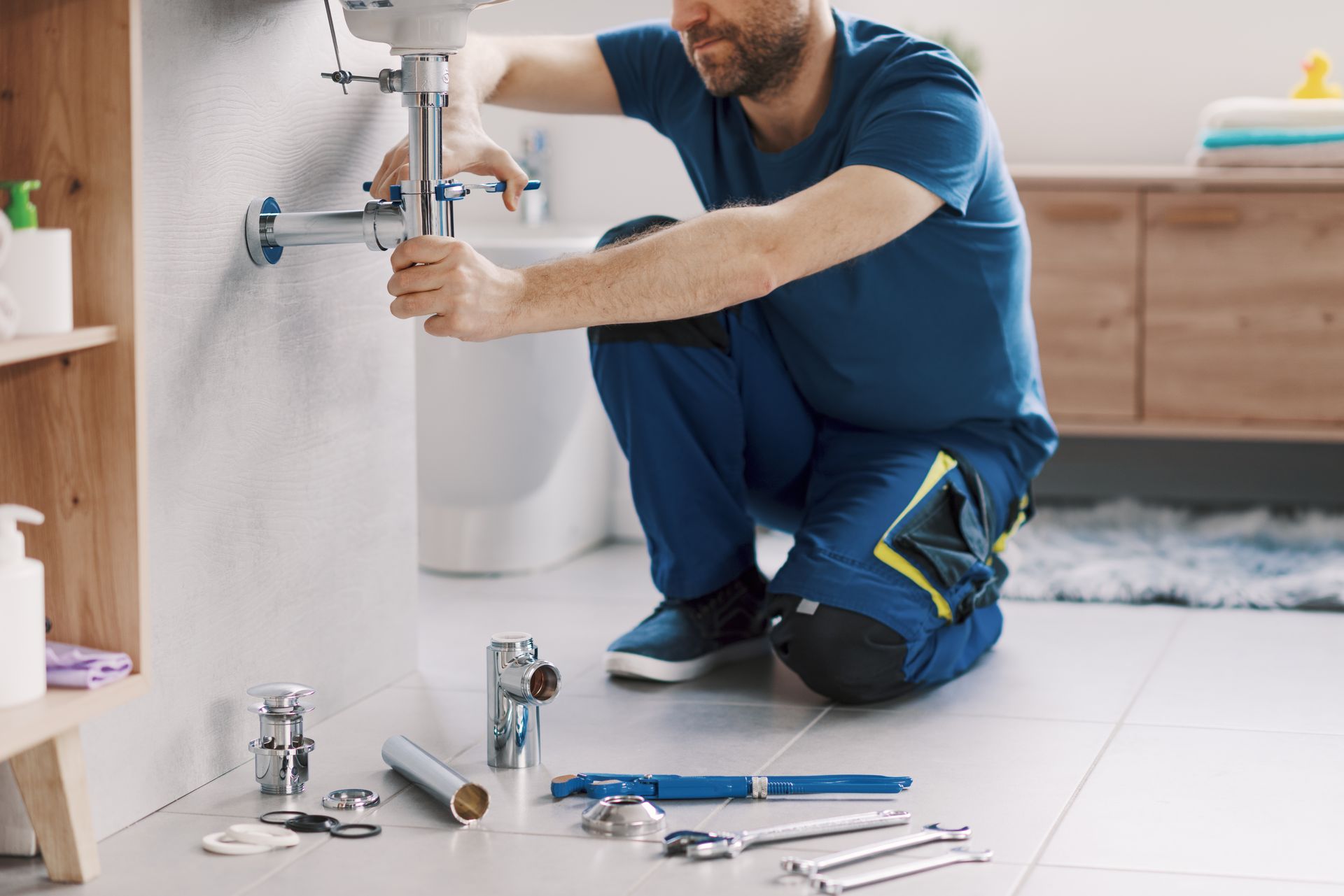 Un plombier agenouillé sous un lavabo, en train de resserrer des tuyaux avec des outils éparpillés sur le sol de la salle de bain.