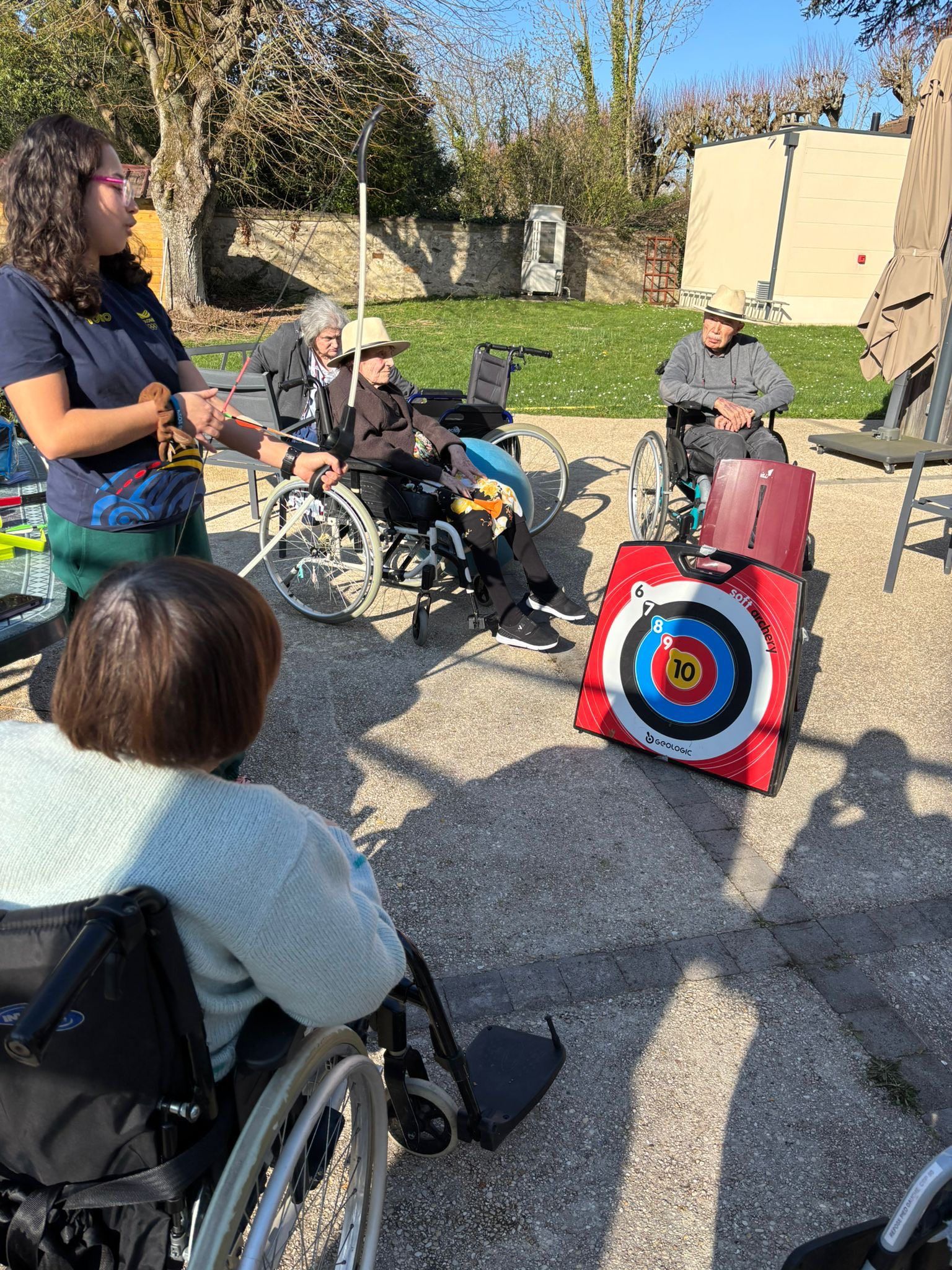 Un groupe de personnes en fauteuil roulant est assis à l'extérieur, face à une cible de tir à l'arc colorée, tandis qu'un membre du personnel leur donne des instructions.