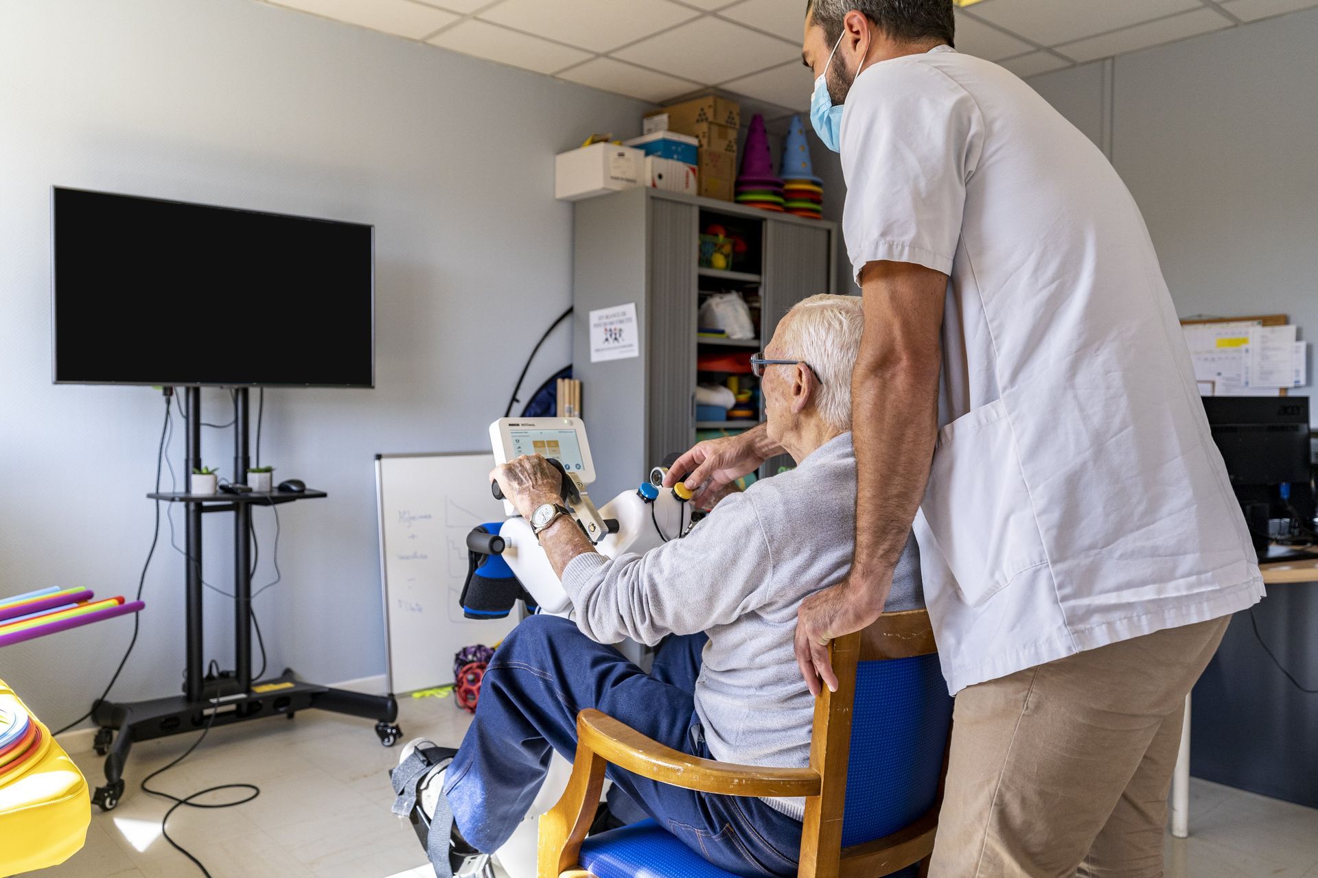 Une aide-soignante se tient près d'un homme âgé qui lit un livre dans une chambre d'hôpital lumineuse, avec une télévision fixée au mur.