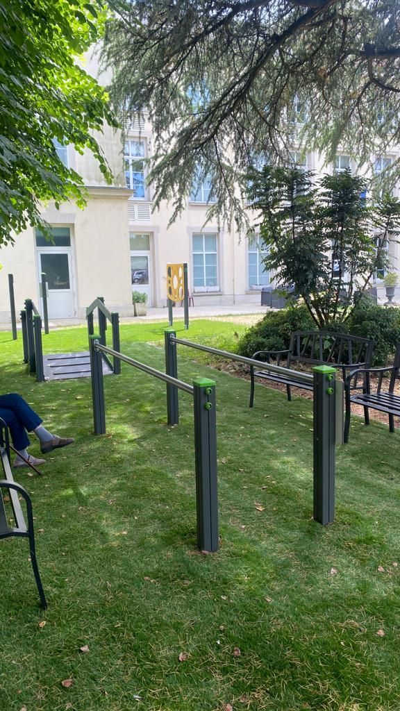 Barres de fitness extérieures dans une cour gazonnée à côté d'un bâtiment blanc, partiellement ombragée par des arbres