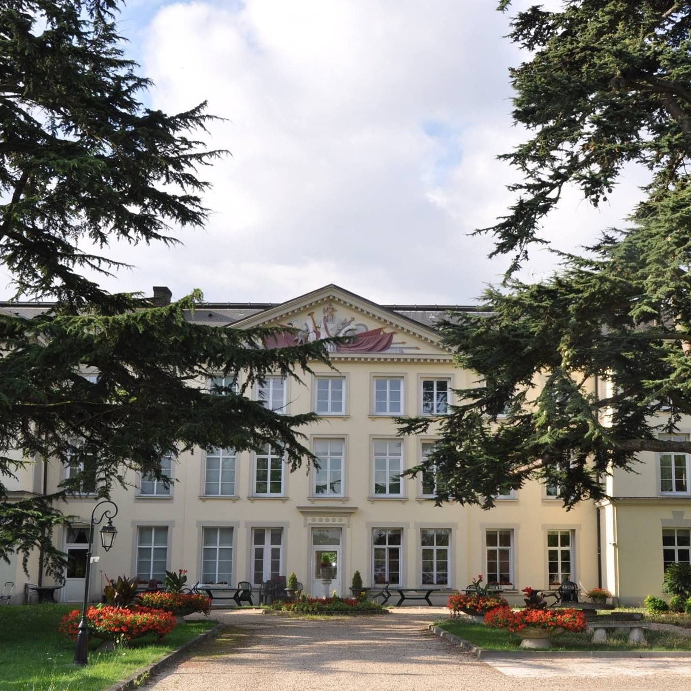 Bâtiment historique couleur crème aux fenêtres blanches, encadré par de grands arbres et des parterres de fleurs sous un ciel nuageux