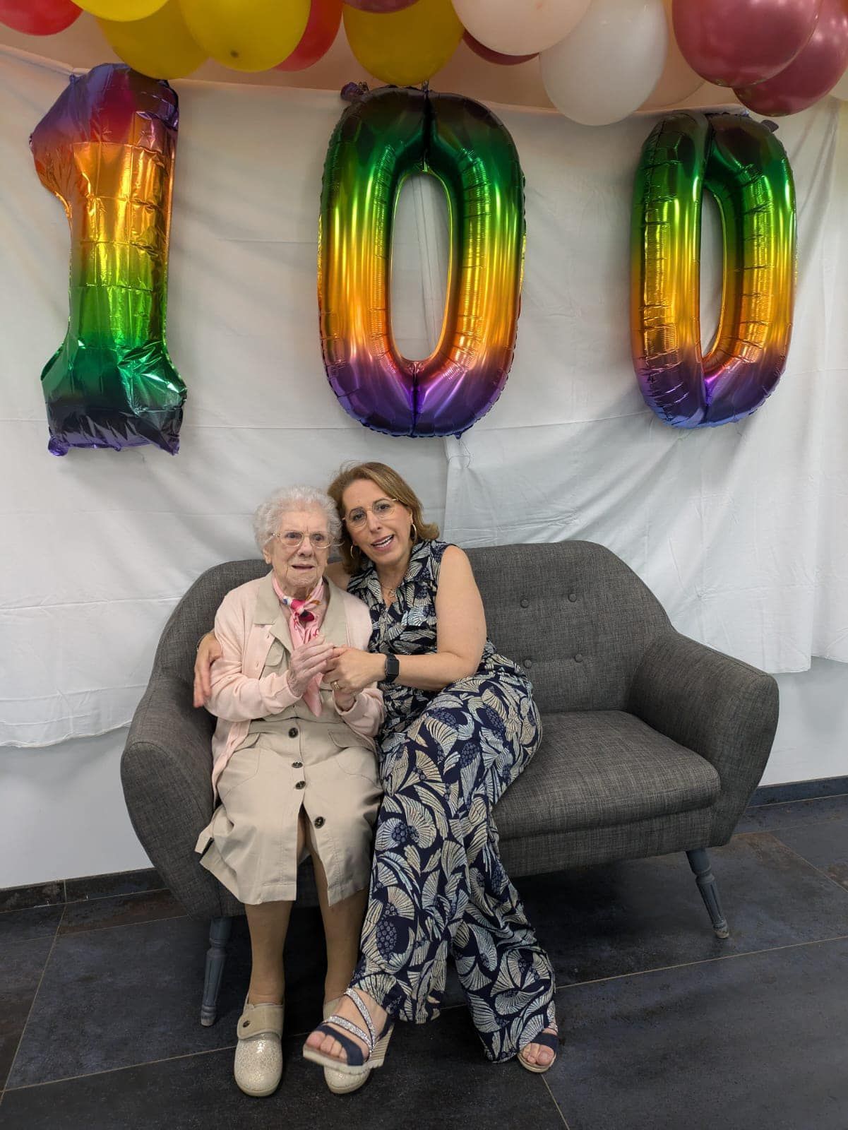 Deux femmes sont assises sur un canapé gris sous des ballons arc-en-ciel formant le chiffre « 100 » et une arche de ballons lors d'une fête.