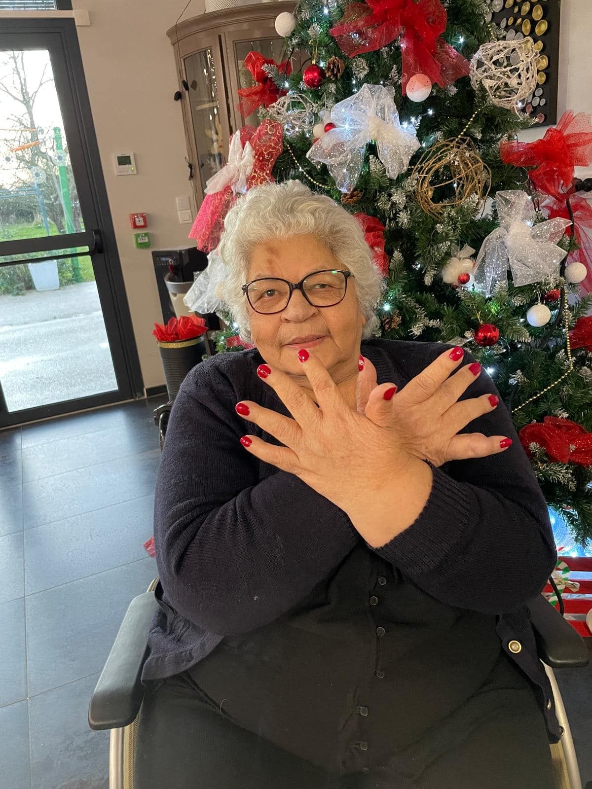 Femme aux cheveux blancs bouclés et aux ongles rouges posant les mains croisées devant un sapin de Noël