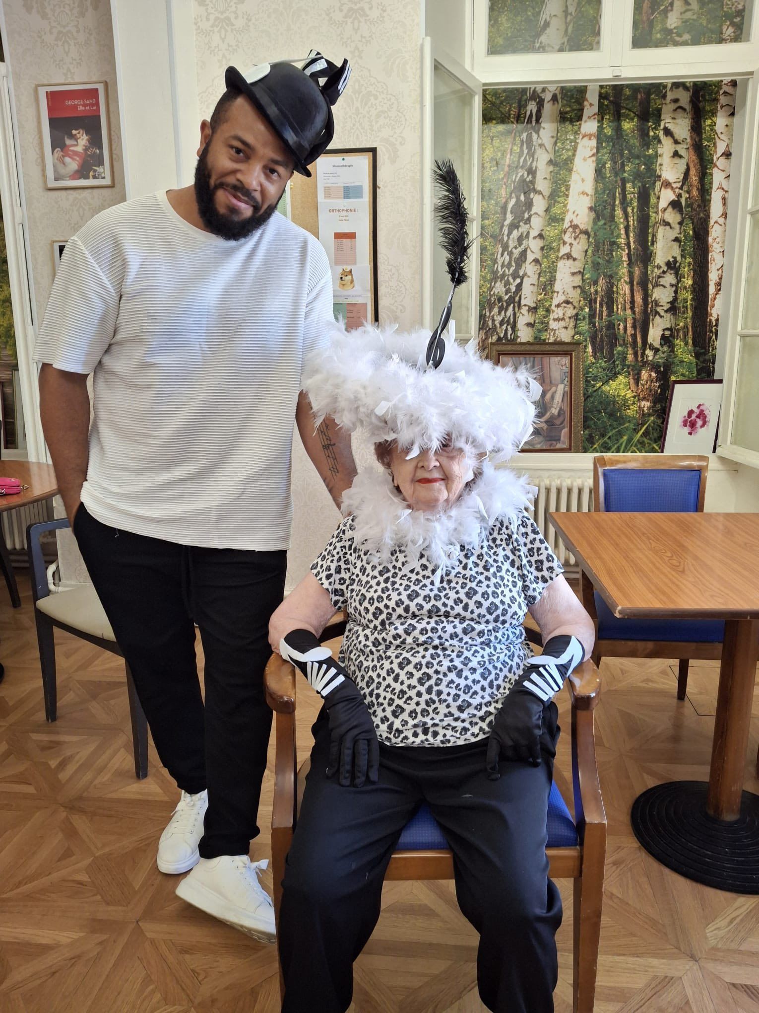 Deux personnes costumées posent à l'intérieur, l'une debout à côté d'une personne assise portant une perruque blanche et un haut à imprimé léopard.