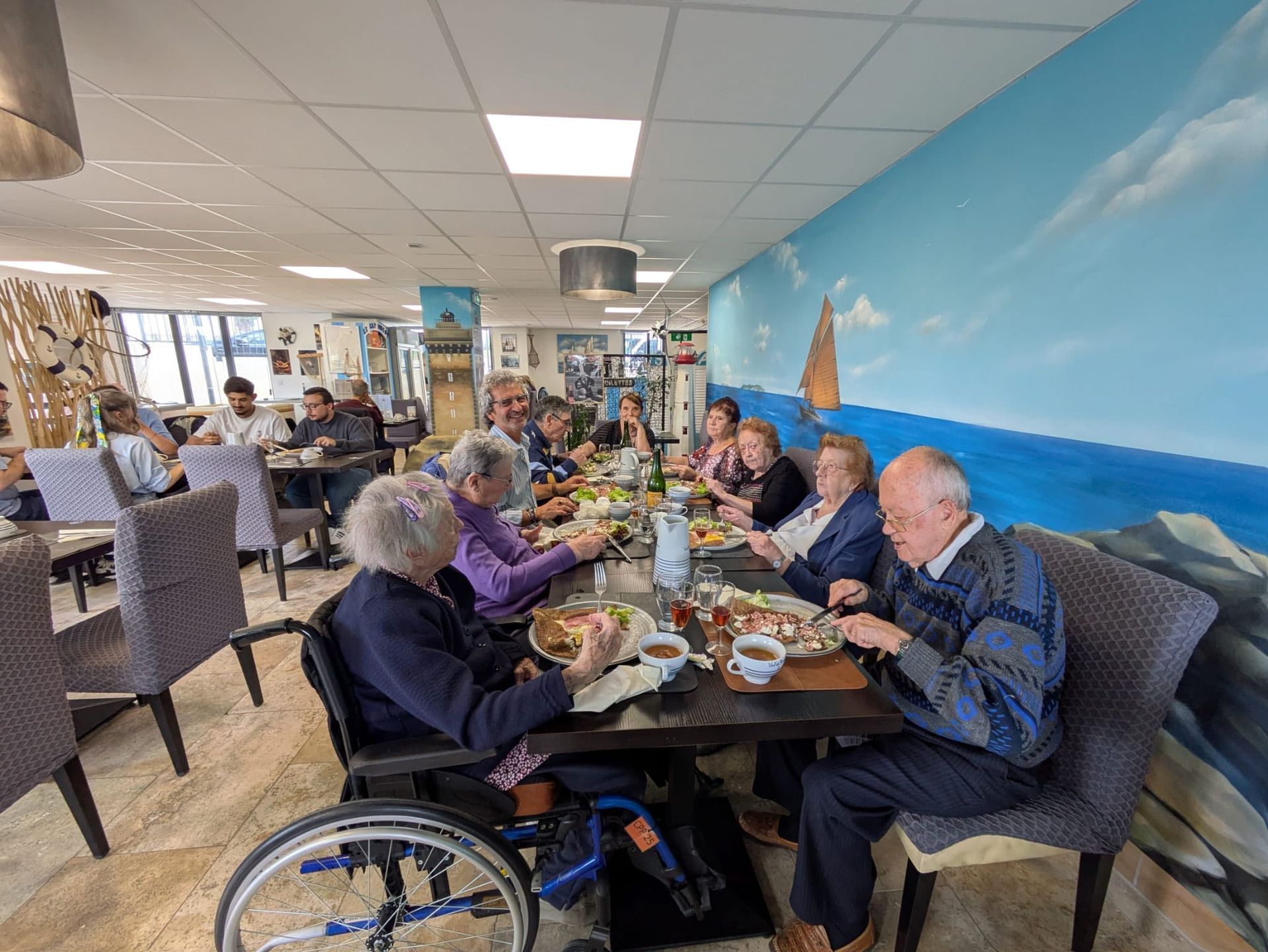 Des clients dînent à une longue table dans un restaurant lumineux en bord de mer, orné d'une fresque et offrant une vue sur l'océan.