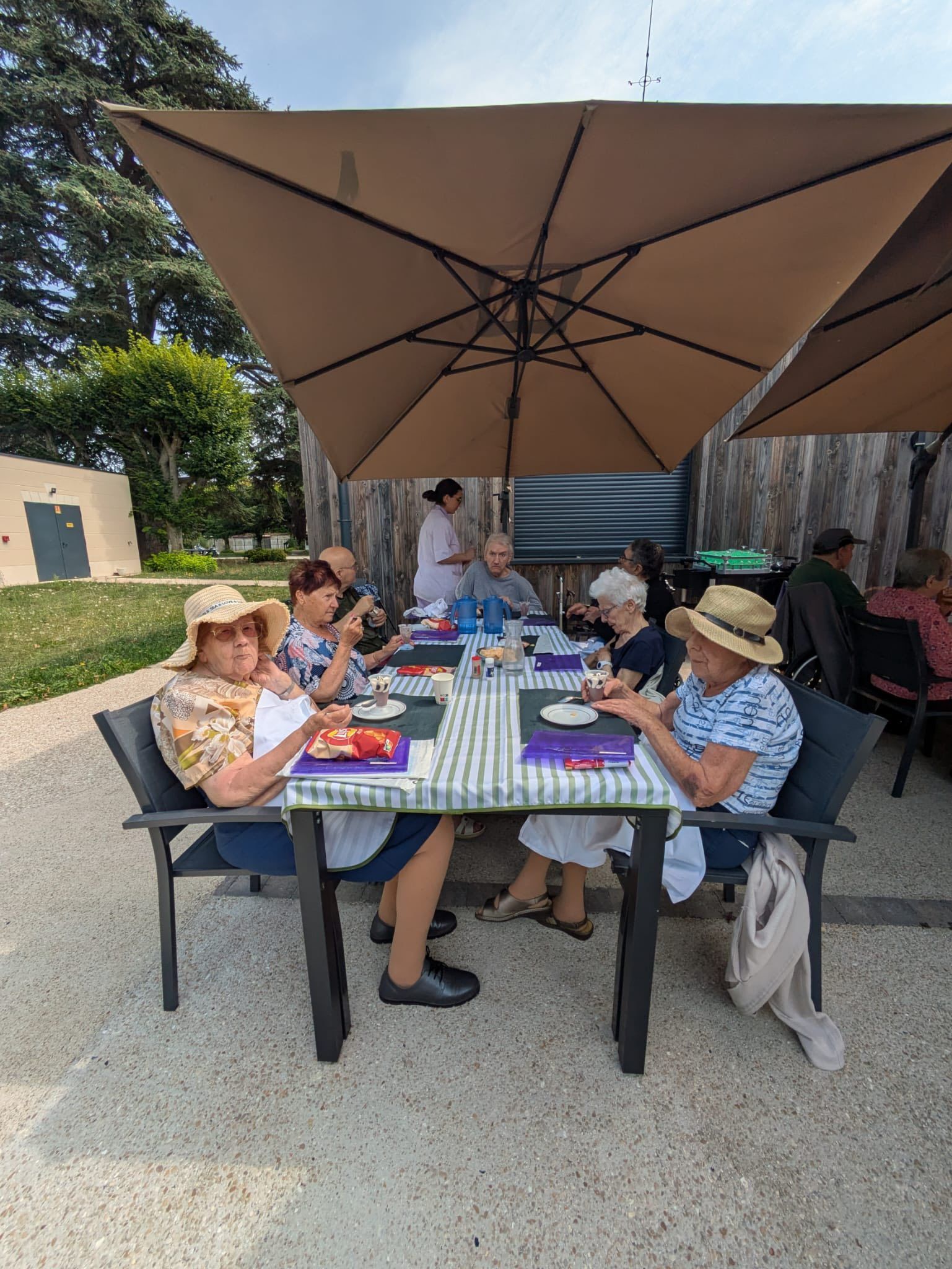 Des personnes assises à une table de pique-nique sous un grand parasol, à l'extérieur, mangent et discutent.