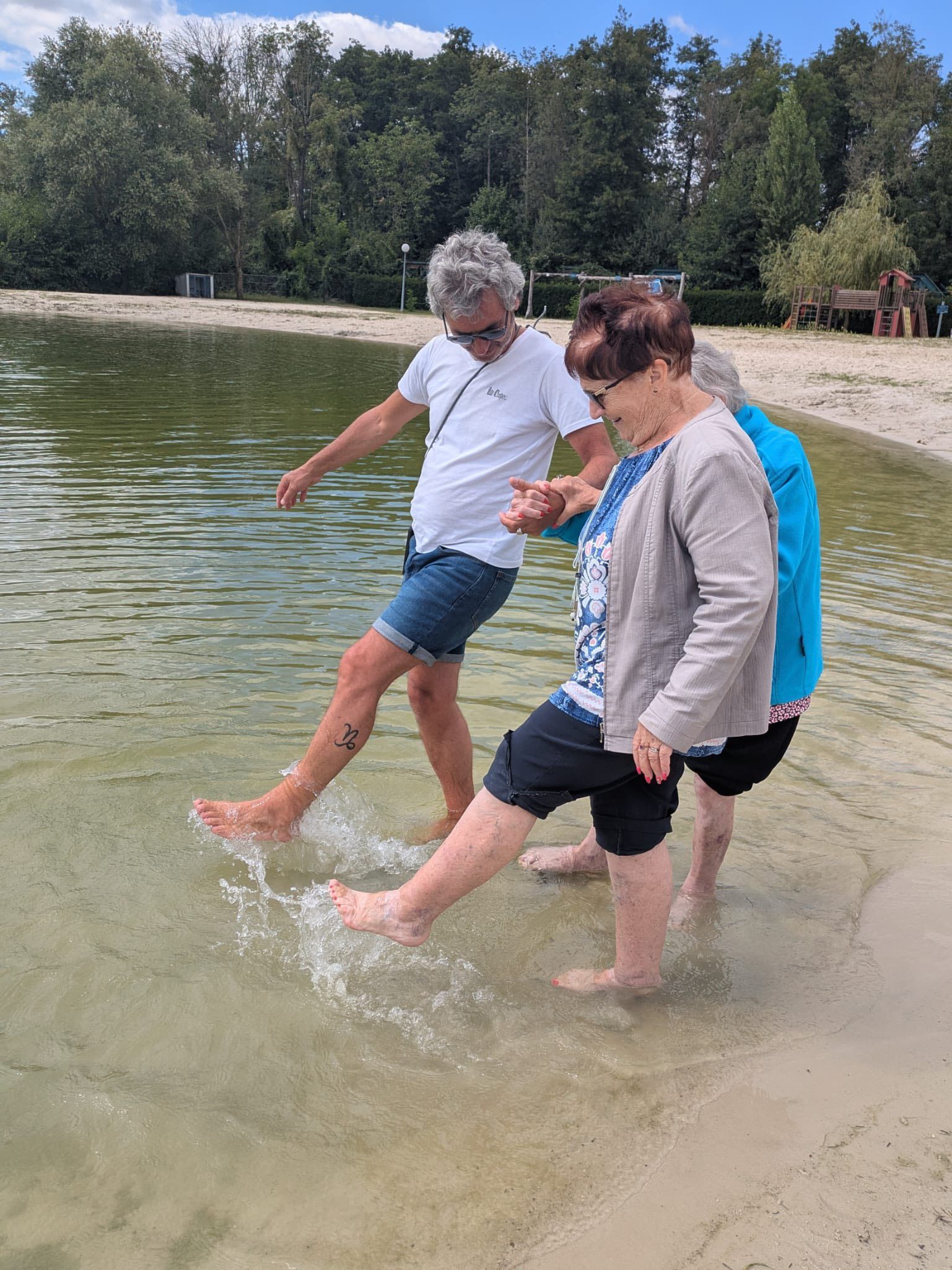 Trois personnes pataugent dans l'eau peu profonde d'un lac, sur une rive sablonneuse, éclaboussant l'eau en marchant.