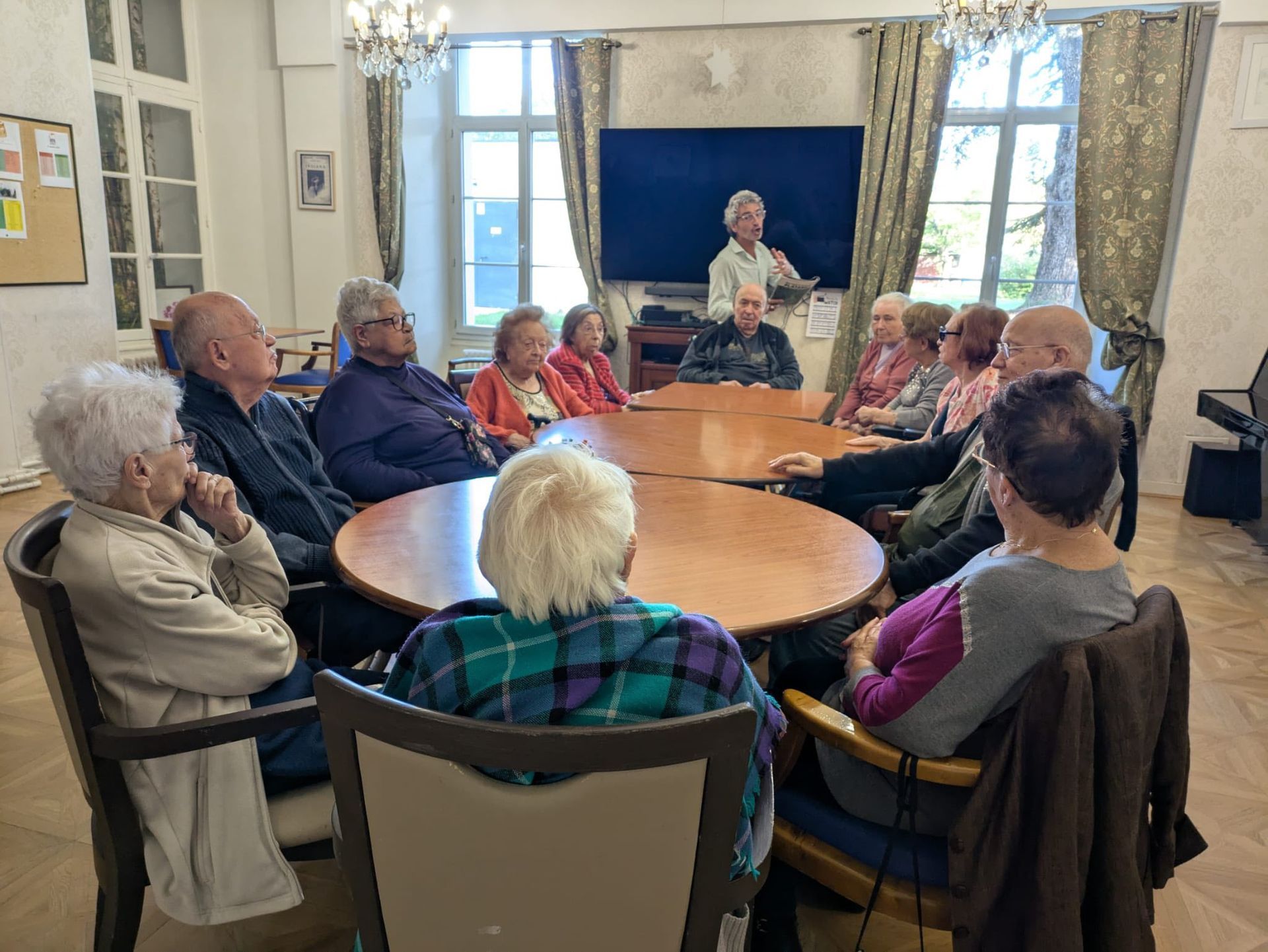 Un groupe de personnes assises autour d'une table ronde dans une pièce lumineuse écoute une femme parler au premier rang.