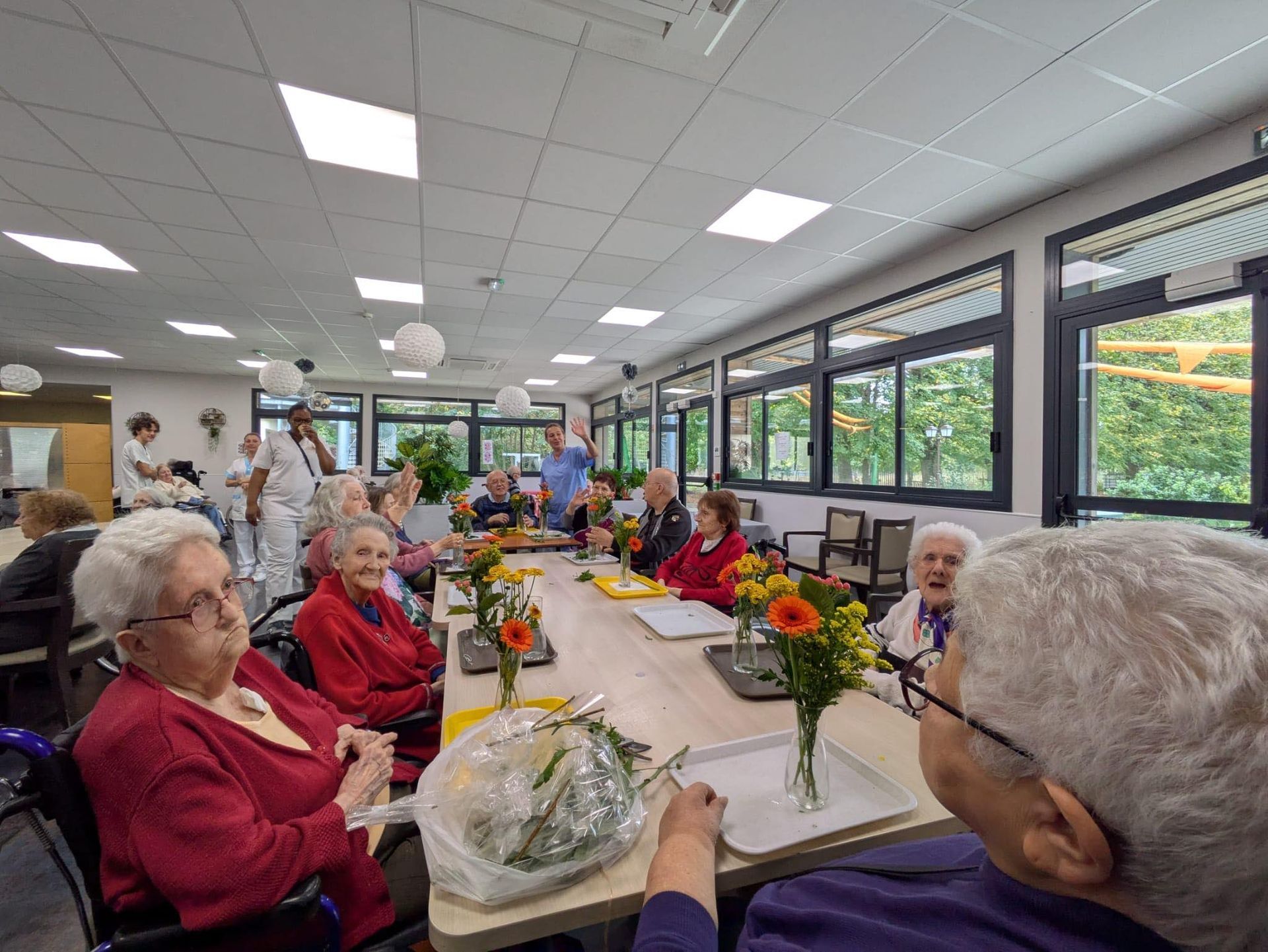Des personnes assises autour d'une longue table dans une salle à manger lumineuse, avec des fleurs et des cadeaux emballés.