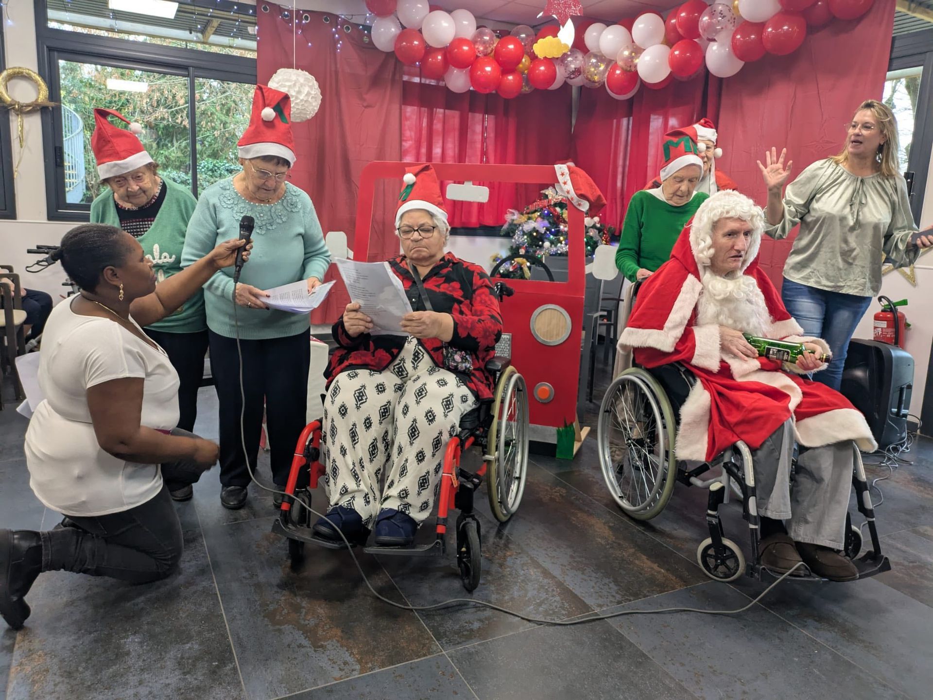 Un groupe de personnes en fauteuil roulant, coiffées de bonnets de Père Noël, pose devant un décor de ballons rouges.