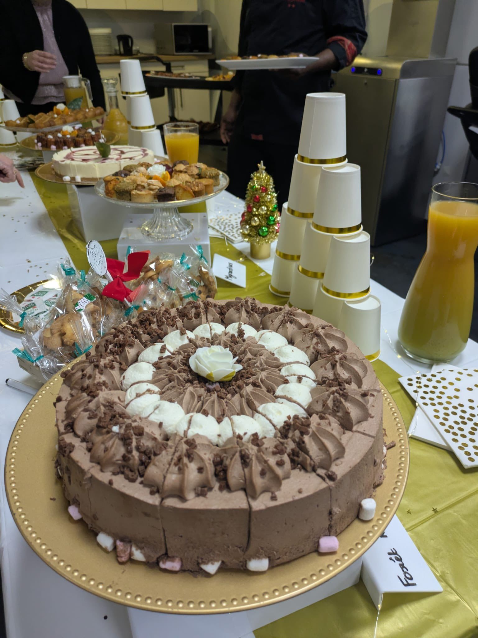 Un gâteau au chocolat sur un plateau doré, devant un buffet garni de friandises de Noël et de tasses blanches empilées en arrière-plan.