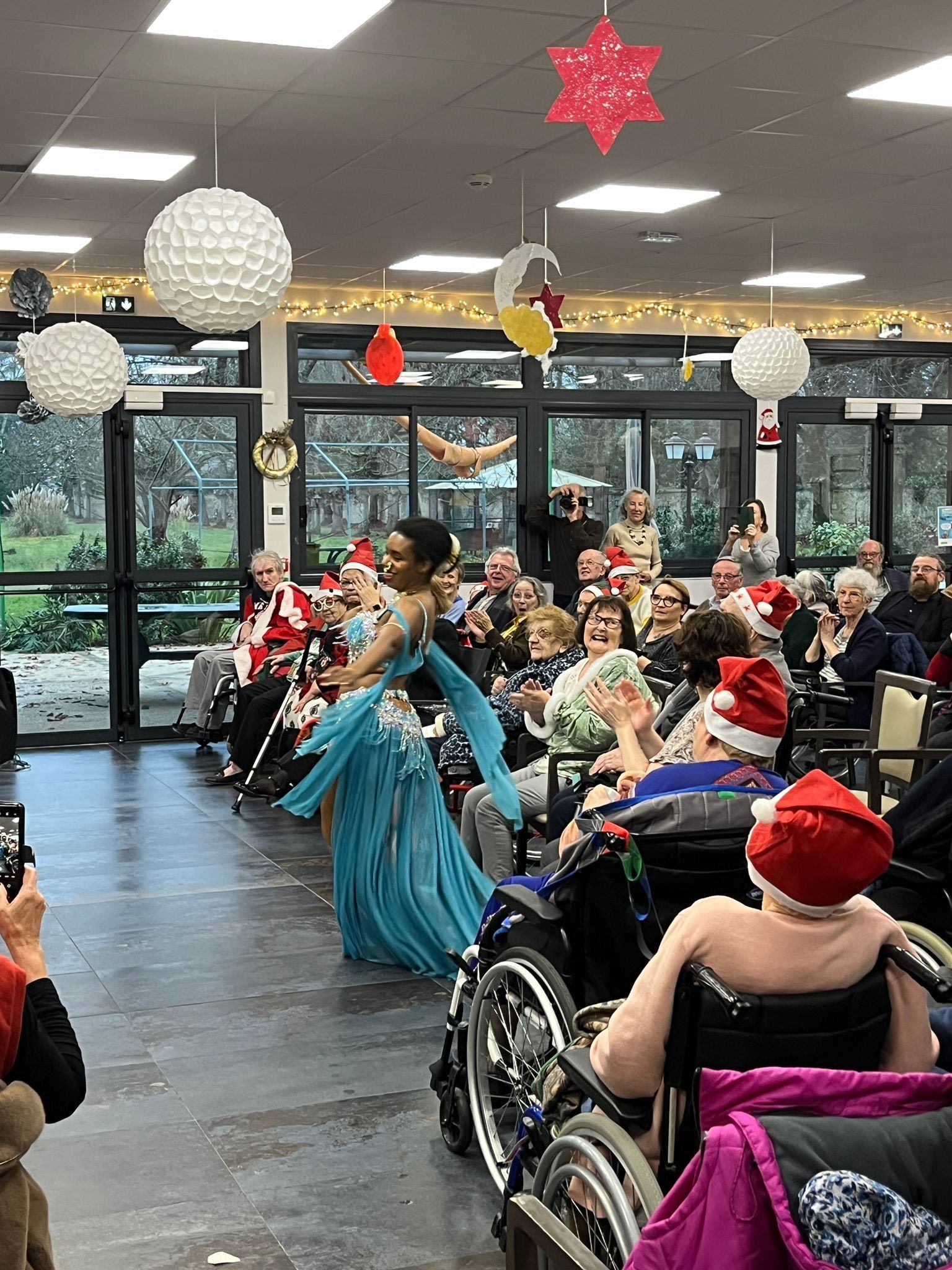 Des danseurs vêtus de bleu et de rouge se produisent dans une salle décorée devant un public assis en fauteuil roulant et sur des chaises.