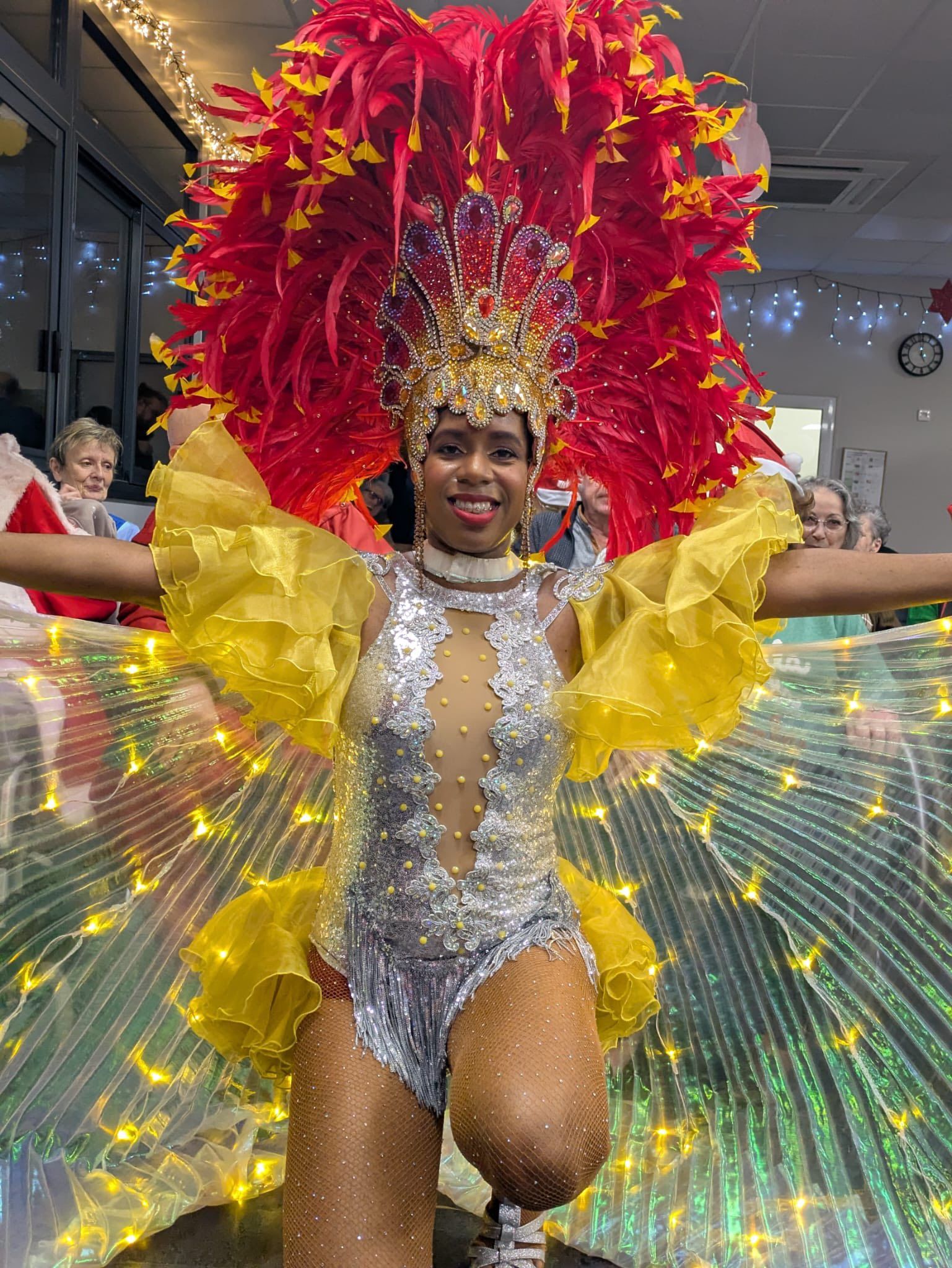 Artiste en costume de carnaval rouge et or, avec des ailes jaunes et un éventail lumineux, posant à l'intérieur, les bras écartés.