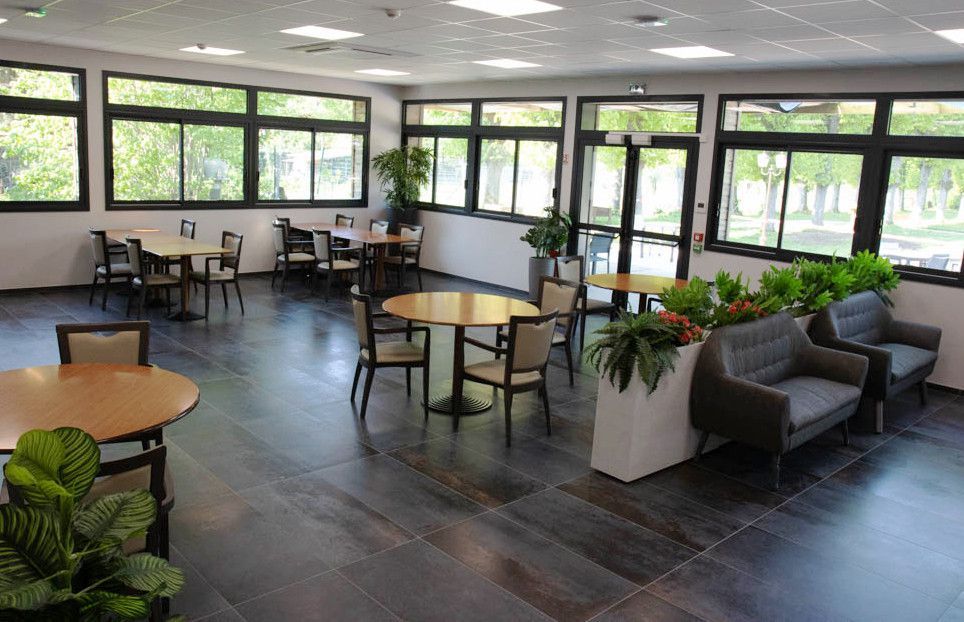 Salon moderne et lumineux avec tables, chaises, canapés et grandes fenêtres donnant sur la verdure.
