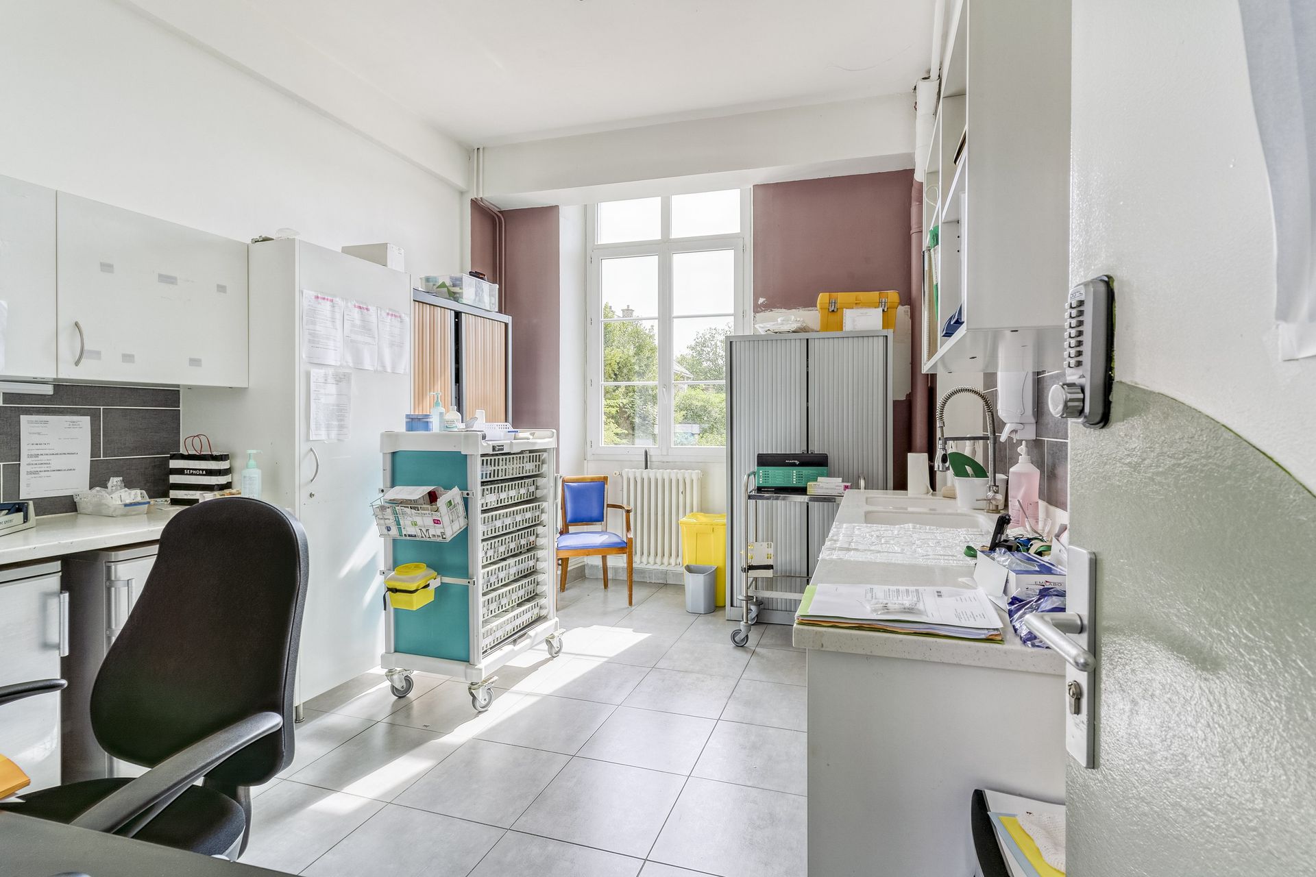Petite salle d'examen médical avec armoires blanches, bureau, chaise, lavabo et fournitures colorées près d'une fenêtre.
