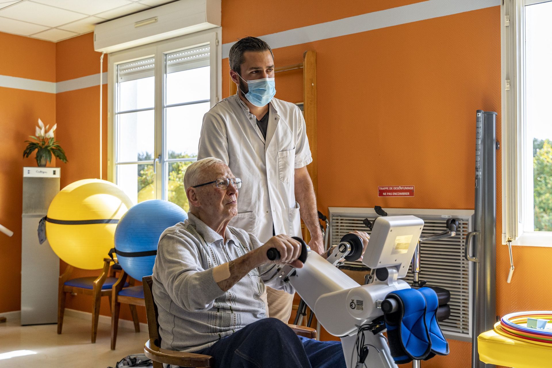Dans une salle de thérapie lumineuse, un thérapeute guide un patient assis à l'aide d'un bras robotisé de rééducation.