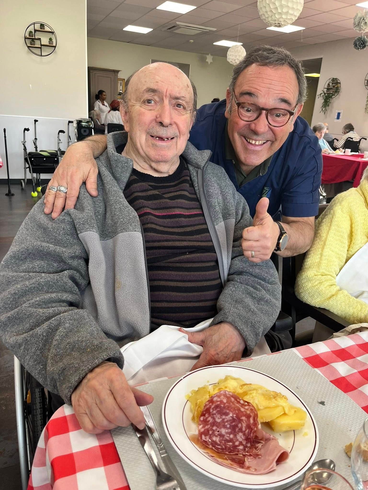 Deux hommes souriants attablés, l'un enlaçant l'autre, une assiette de nourriture devant eux.