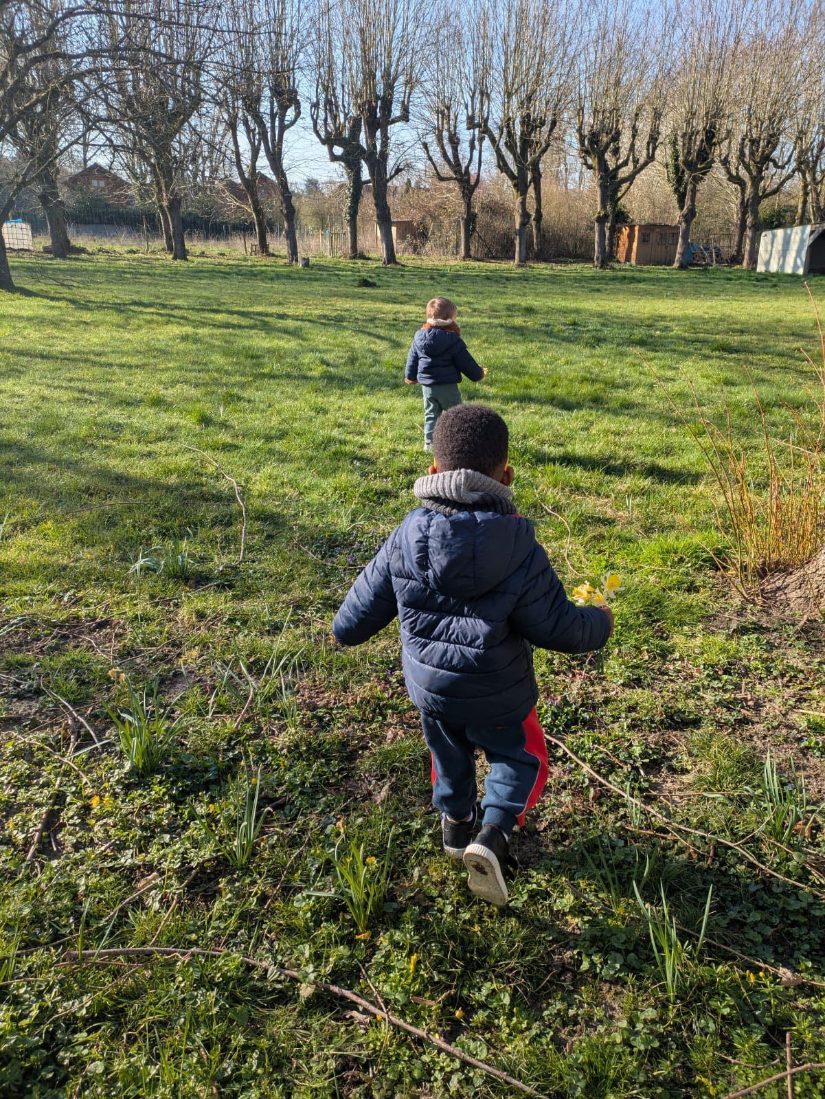 Deux enfants en manteaux d'hiver courent à travers un champ herbeux vers des arbres dénudés par une journée ensoleillée.