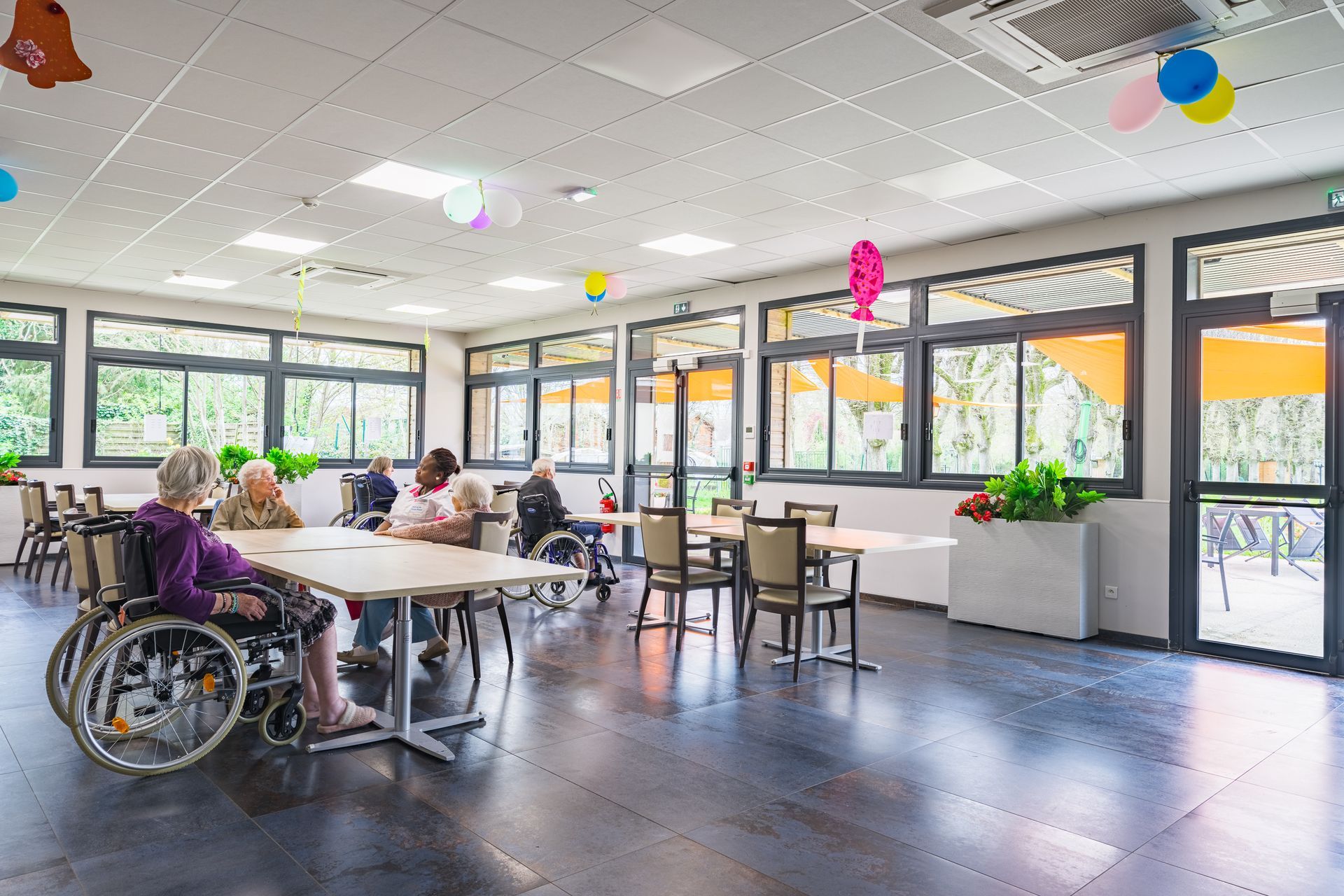 Des personnes assises à des tables dans une salle communautaire lumineuse, avec une personne en fauteuil roulant au premier plan.