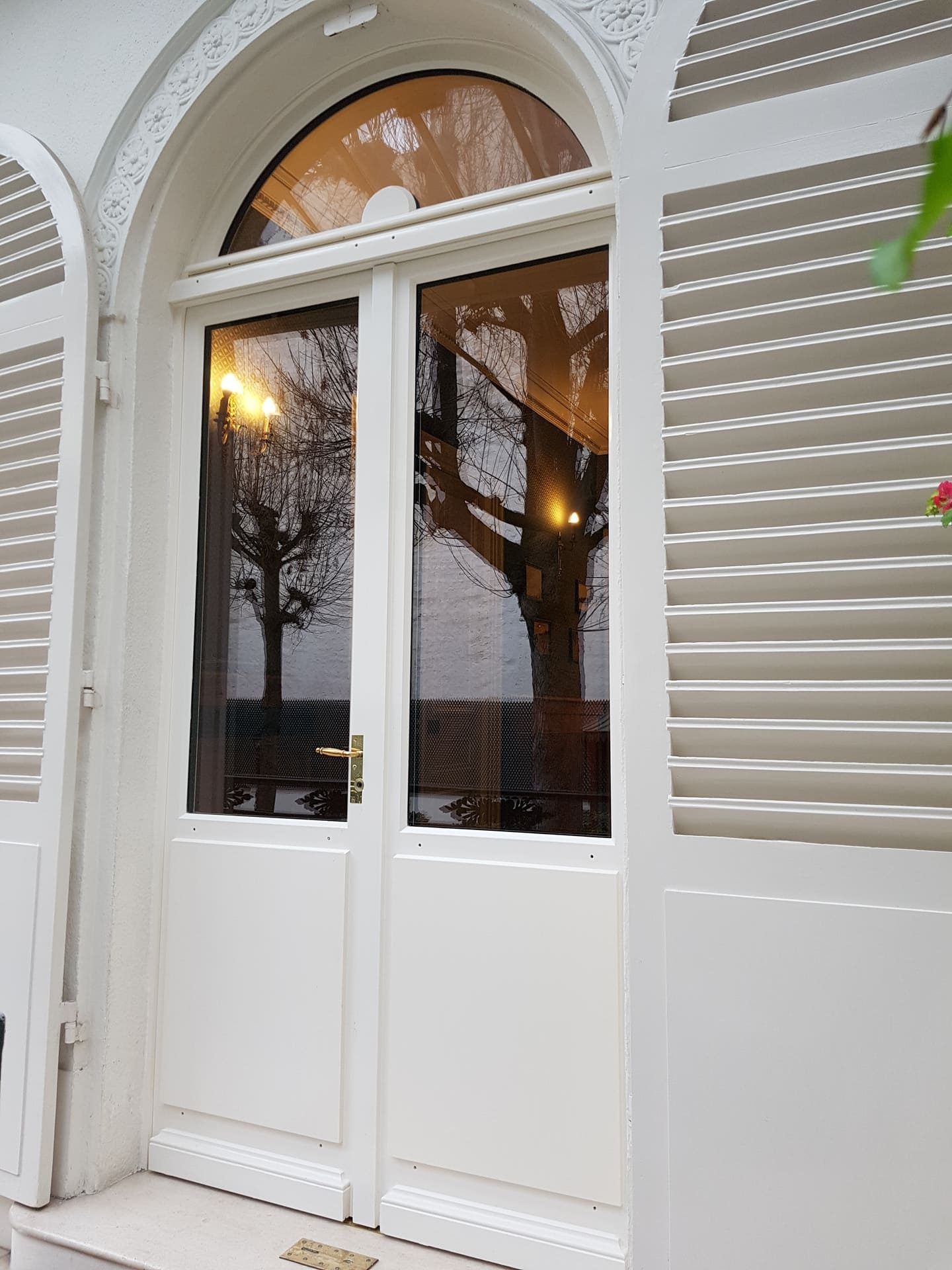 Une double porte cintrée blanche à panneaux de verre, adossée à un mur blanc assorti, orné de volets latéraux.