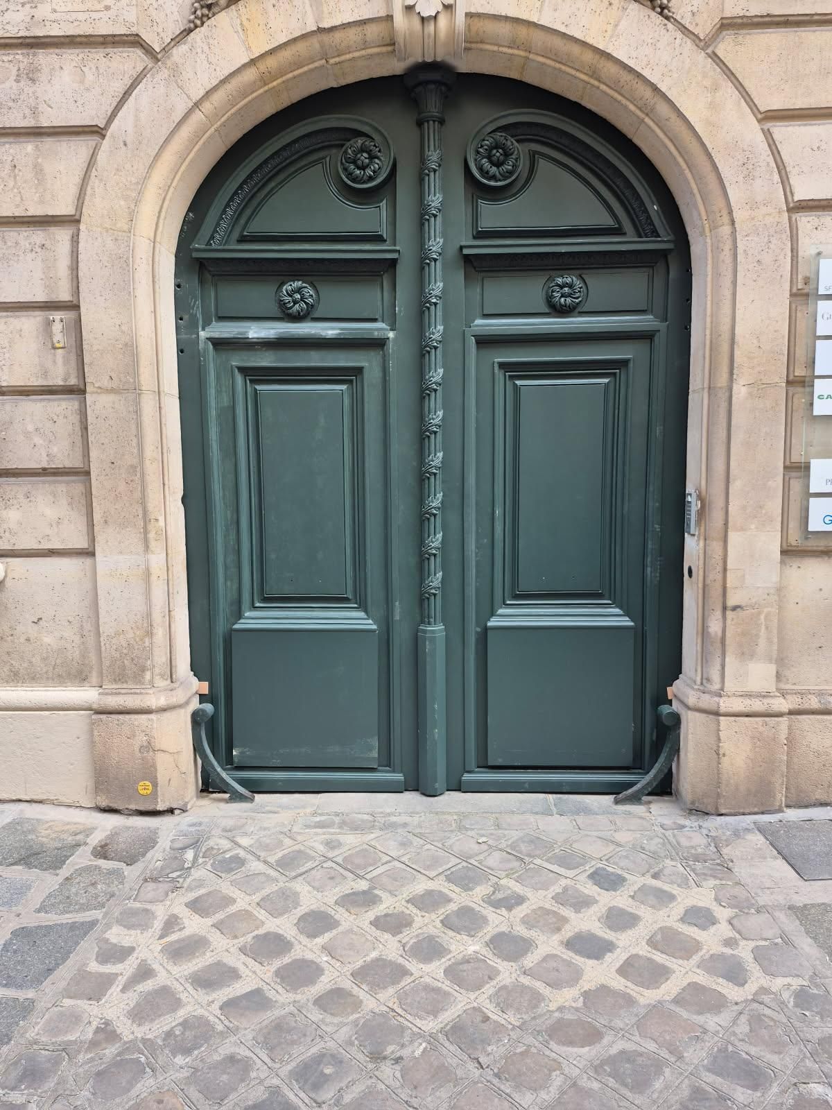 Double porte en bois vert ornée de détails décoratifs, dans une entrée cintrée en pierre, sur une rue pavée.