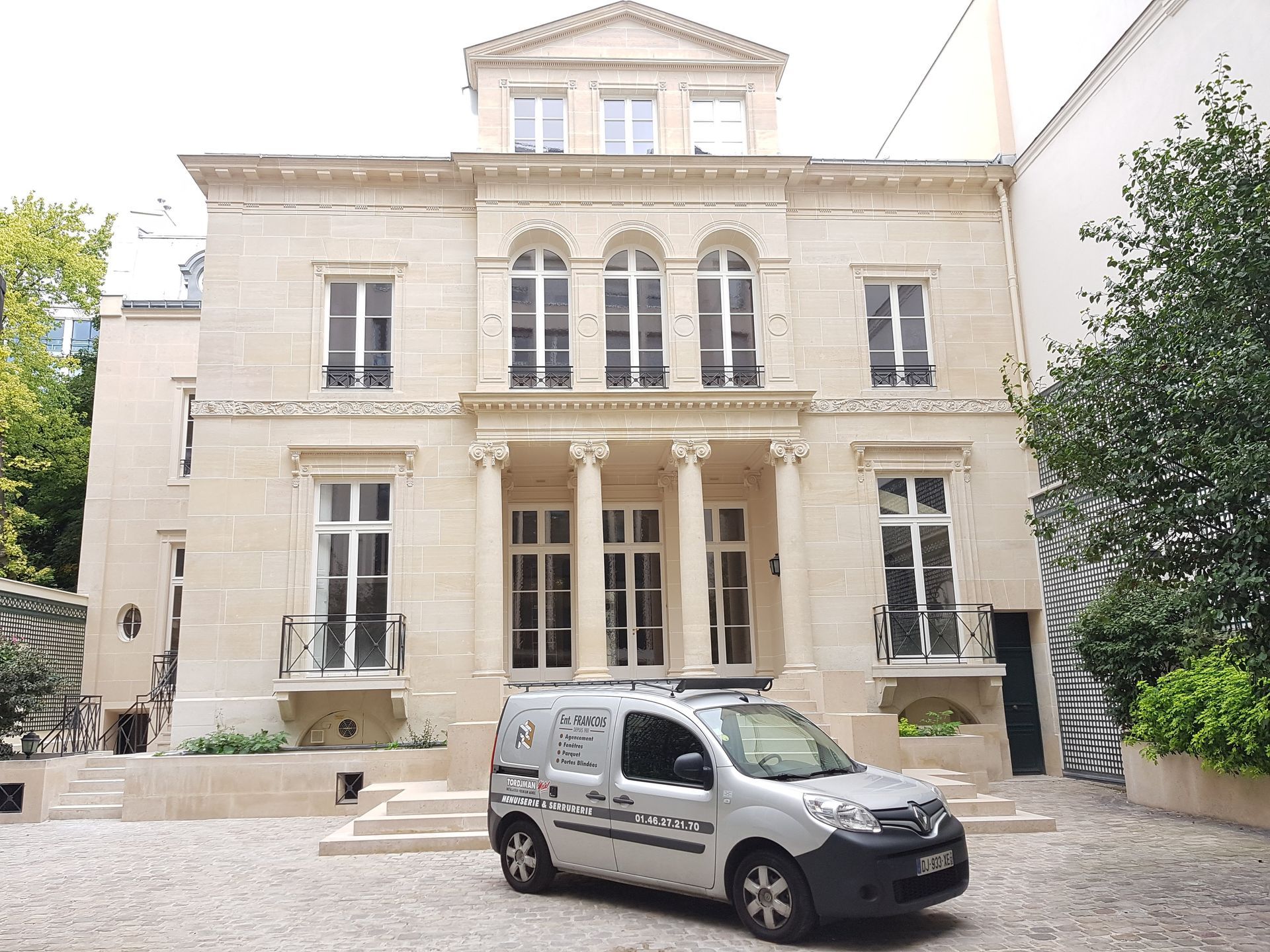 Une camionnette argentée est garée devant un bâtiment de trois étages en pierre claire, avec un portique central à colonnes.