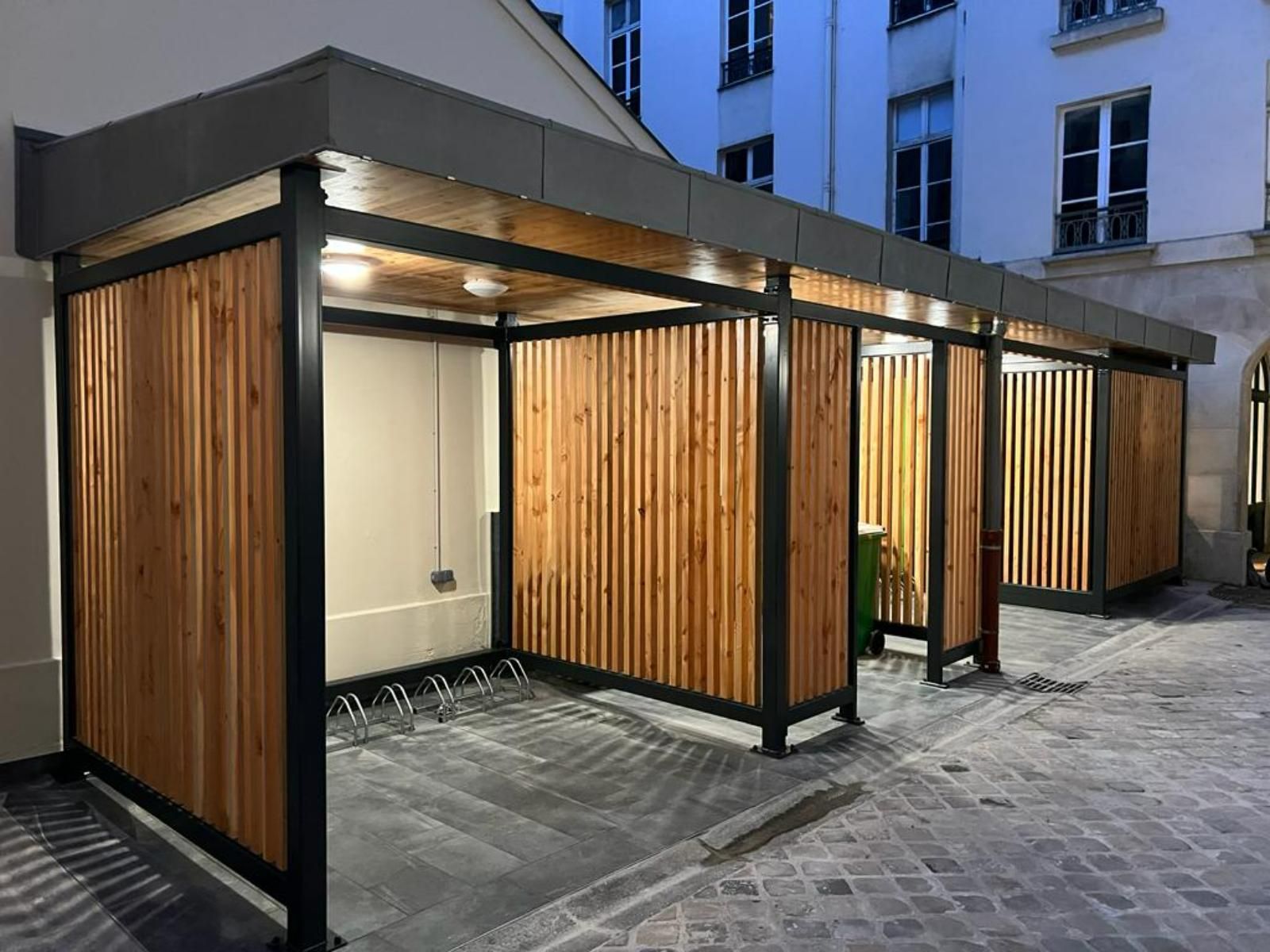 Abri à vélos avec lattes de bois et structure en métal noir, éclairé par des lampes suspendues, dans une rue pavée.