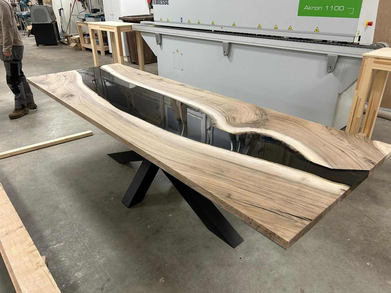 Table en bois avec rivière en résine noire, reposant sur un piètement en métal noir, dans un environnement d'atelier.