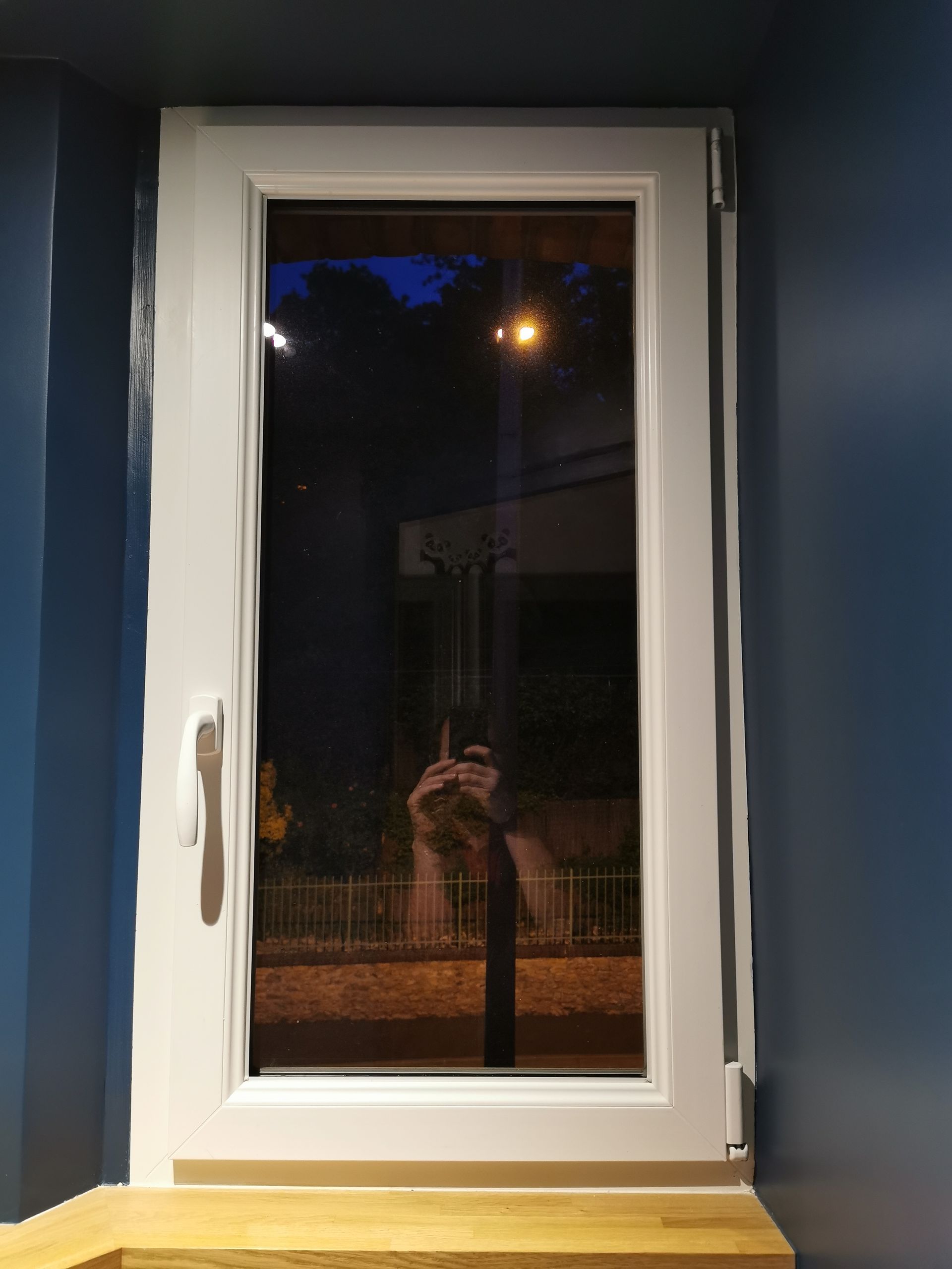 Une fenêtre à cadre blanc la nuit, avec un encadrement mural bleu foncé et un appui de fenêtre en bois en dessous.