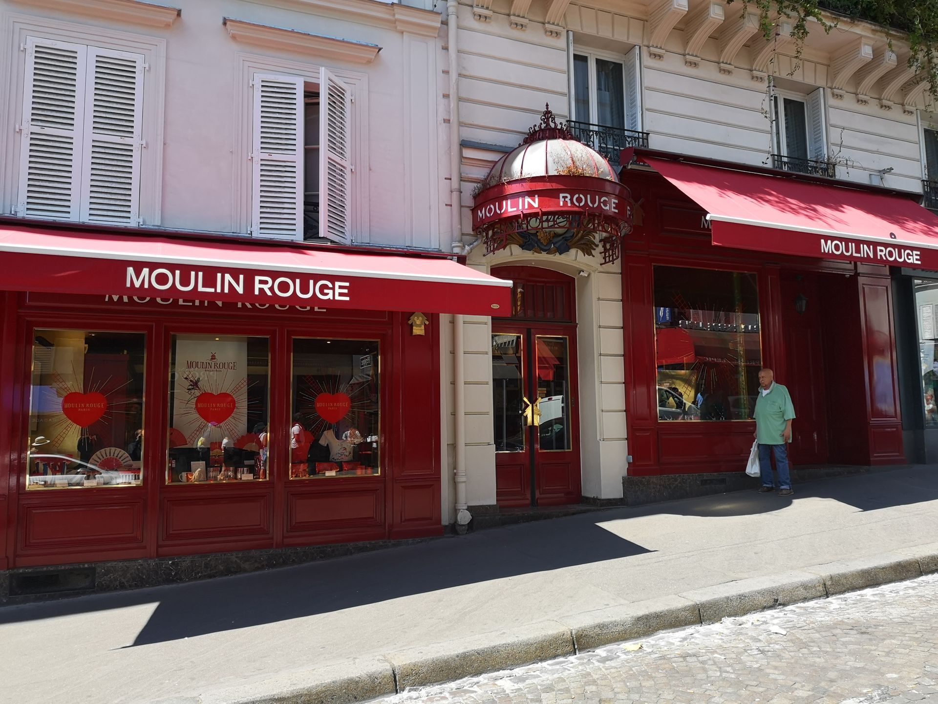 Une boutique à façade rouge portant l'inscription « Moulin Rouge », avec des volets blancs et une personne debout près de l'entrée, dans une rue pavée.