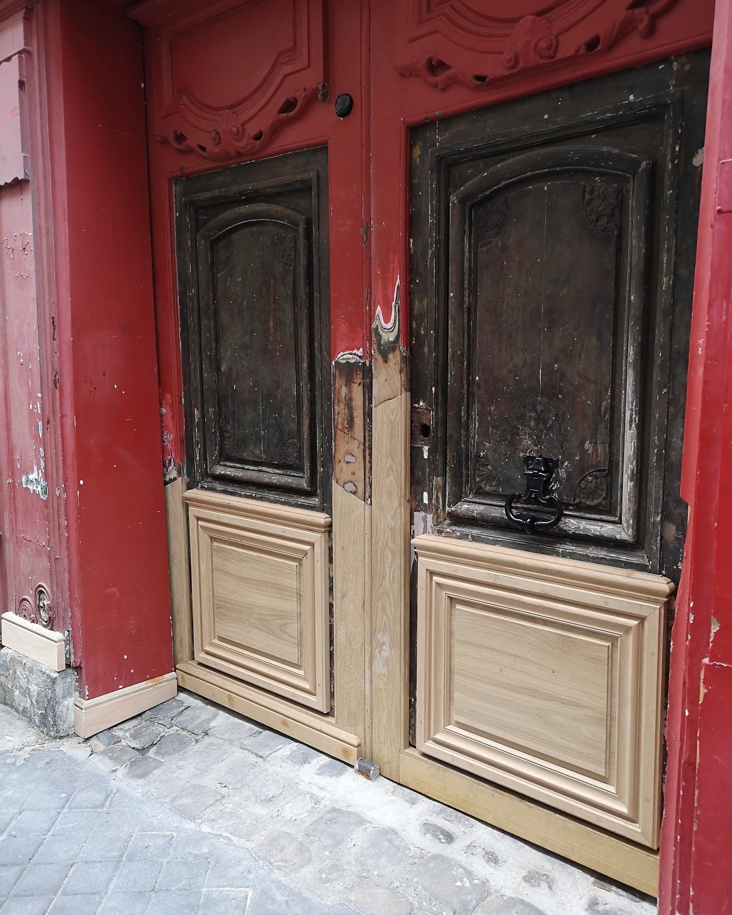 Une double porte en bois à la peinture rouge écaillée, partiellement rénovée avec des panneaux inférieurs en bois clair, installée dans une allée en pierre.