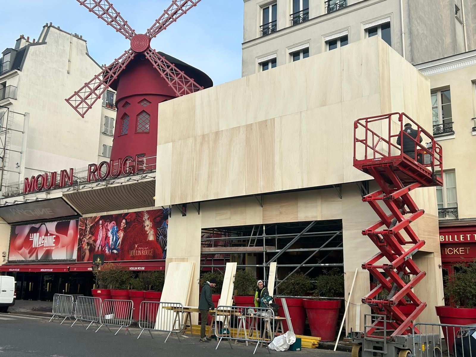 Un moulin à vent rouge vif, le Moulin Rouge, se dresse derrière un bâtiment partiellement masqué par des palissades de chantier en bois.