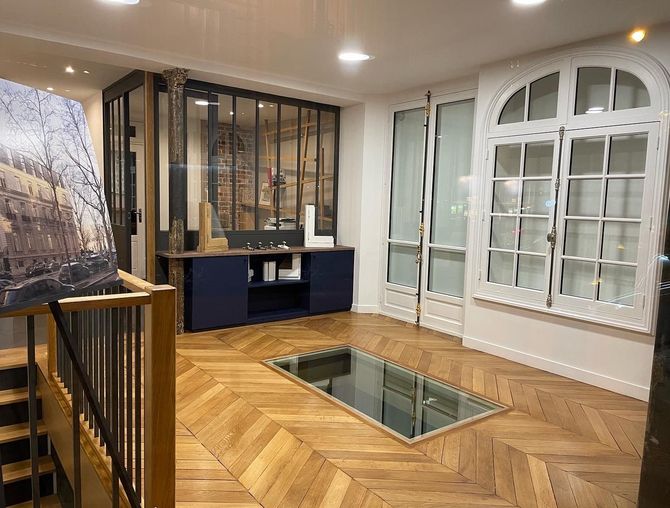 Un showroom moderne avec parquet clair à chevrons, un insert en verre au sol, des armoires bleu foncé et des fenêtres cintrées.