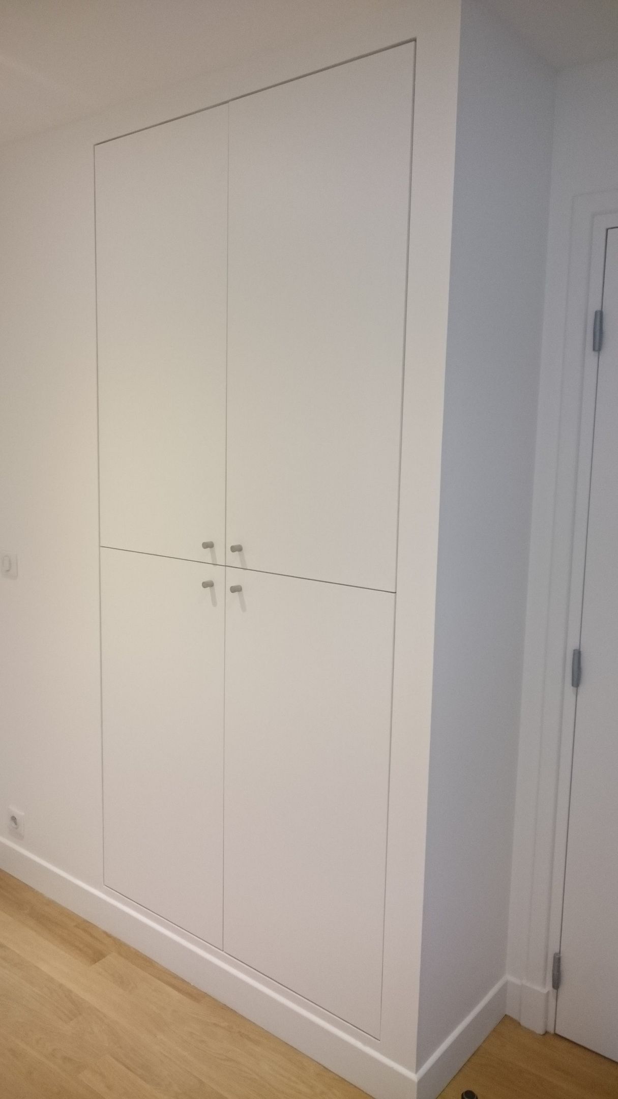 Une grande armoire encastrée blanche, allant du sol au plafond, avec quatre portes et deux jeux de poignées argentées, installée dans une pièce avec parquet.