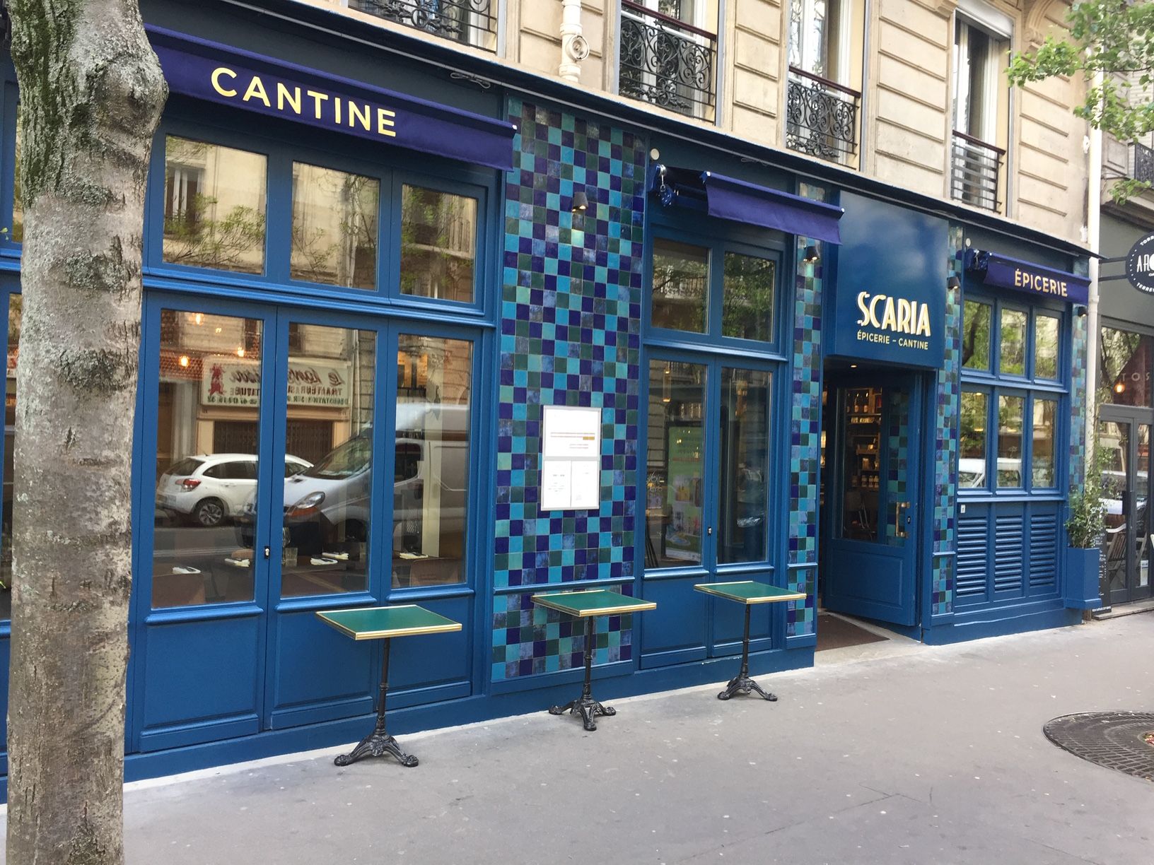 La façade bleue du restaurant Scaria, avec ses petites tables en terrasse sur le trottoir, devant un arbre et des bâtiments urbains.