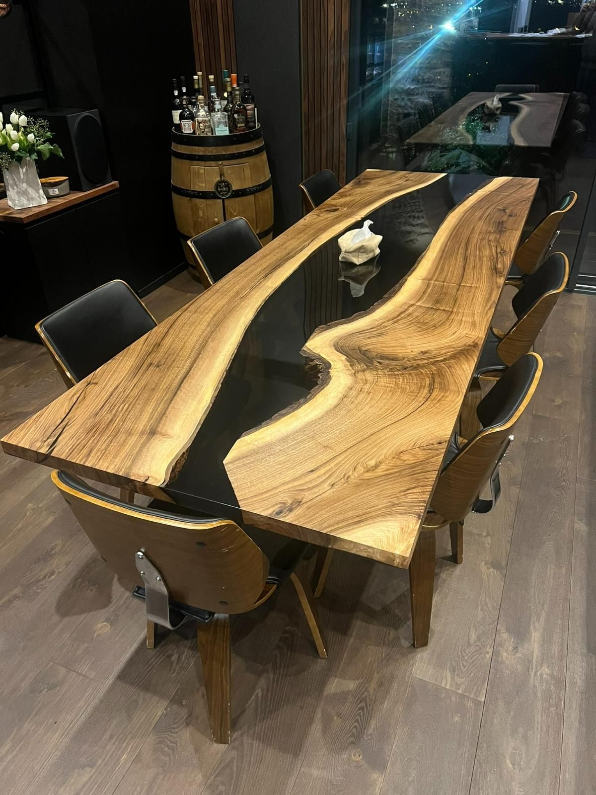 Grande table en bois avec un décor de rivière en époxy noir, entourée de chaises noires et en bois, à l'intérieur.
