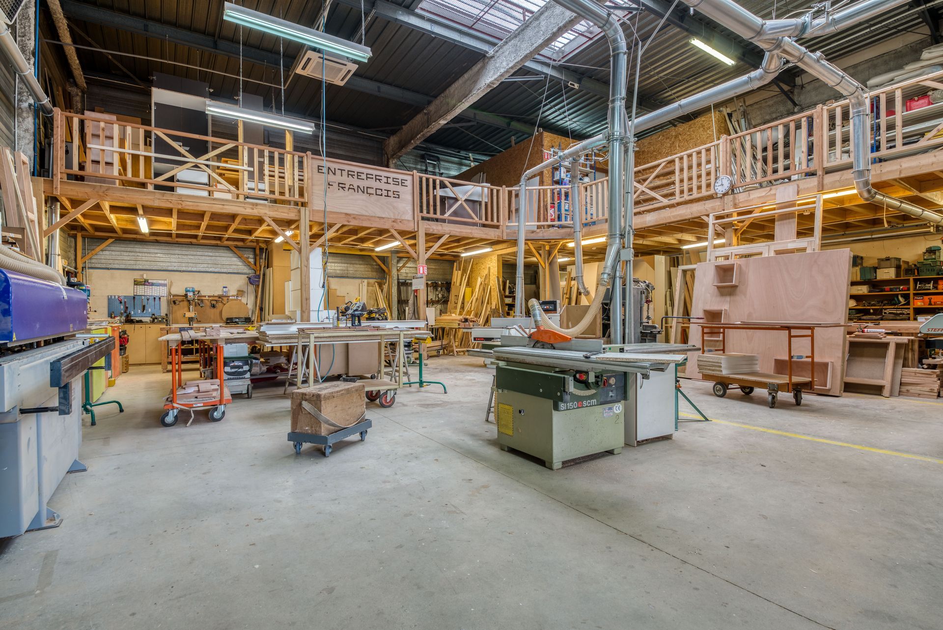 Un vaste atelier de menuiserie avec des établis, des outils électriques stationnaires et une mezzanine en bois.