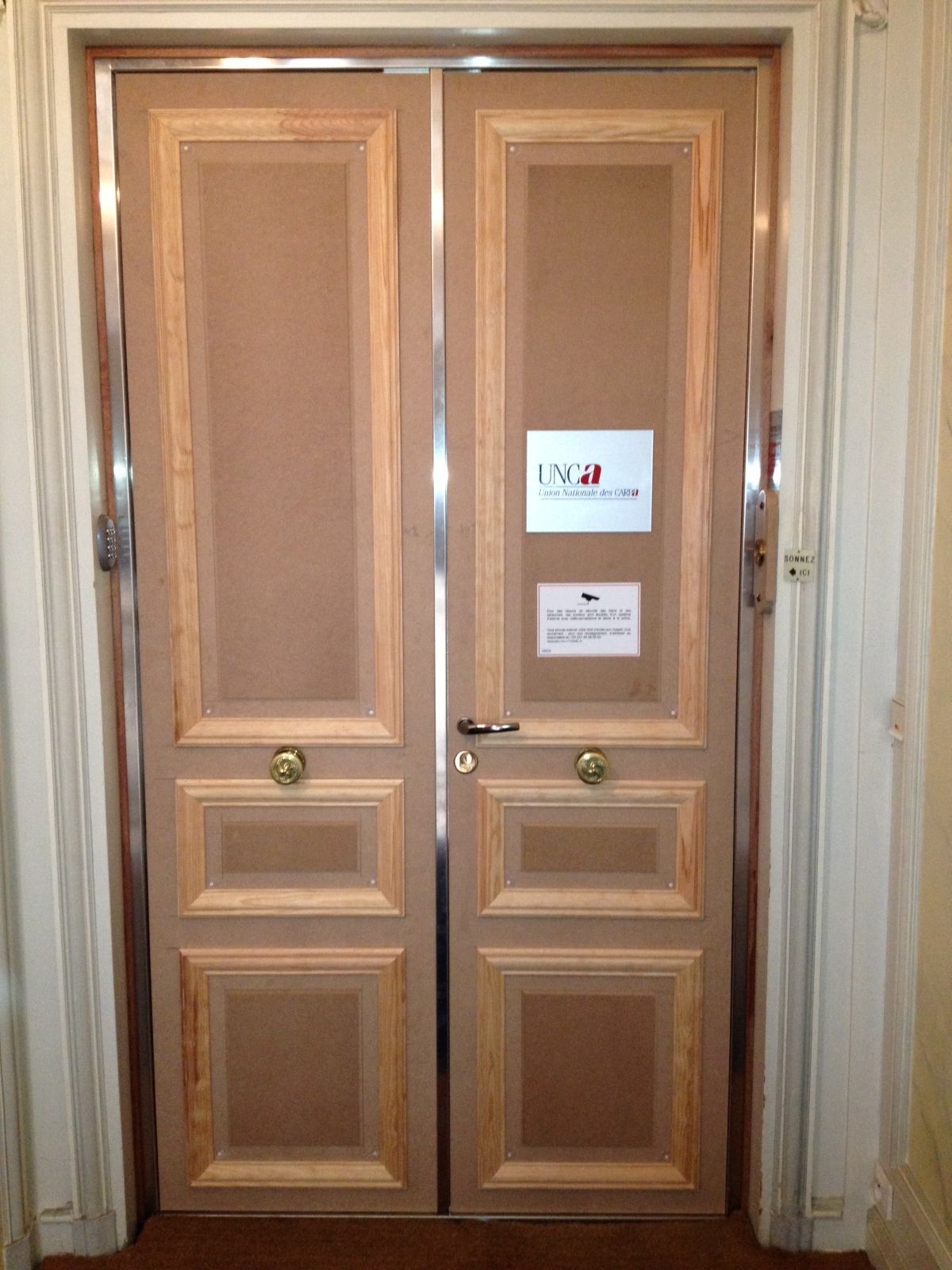 Deux portes doubles en bois clair, encadrées de blanc, avec un panneau central orné de détails et deux petits panneaux d'information.