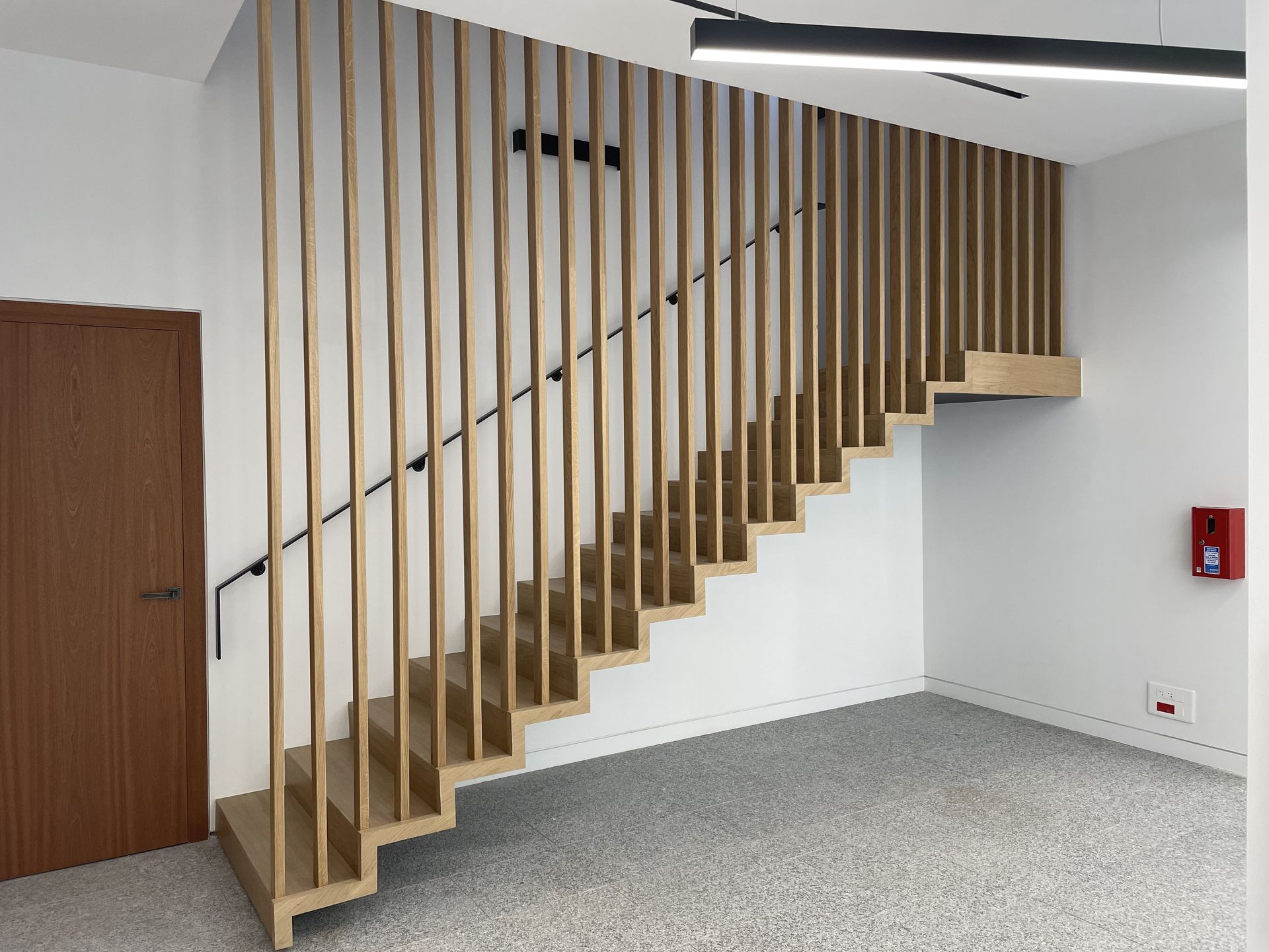 Un escalier en bois avec des rampes à lattes verticales dans une pièce moderne avec un sol gris moucheté et un mur blanc.