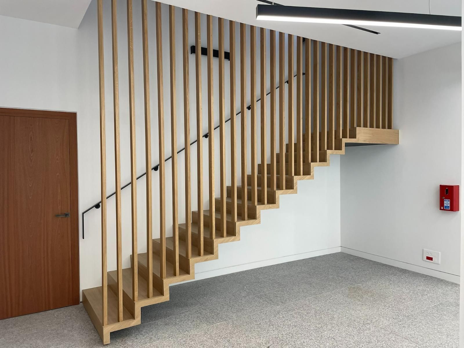 Escalier en bois avec rampe à lattes. Murs blancs, porte en bois et sol moucheté.