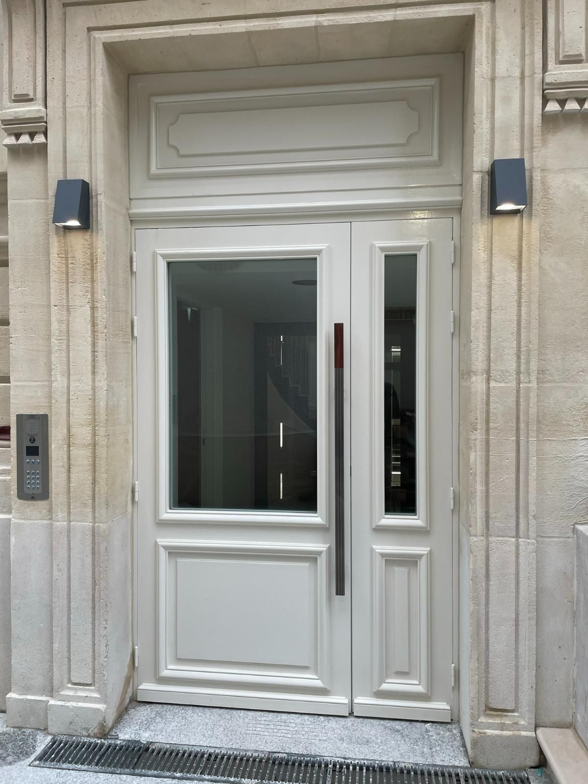 Entrée d'un immeuble blanc avec porte vitrée et imposte, poignée argentée. Deux appliques noires sur une façade en pierre.