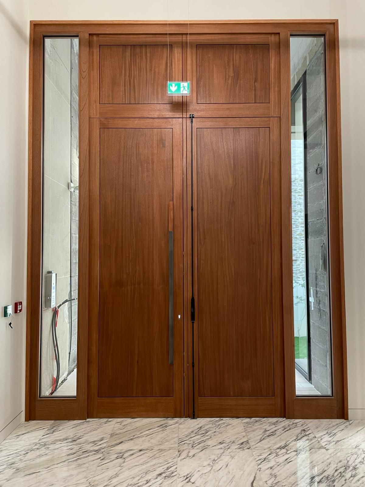 Double porte en bois avec panneaux latéraux vitrés, panneau de sortie au-dessus. Sol en marbre clair.
