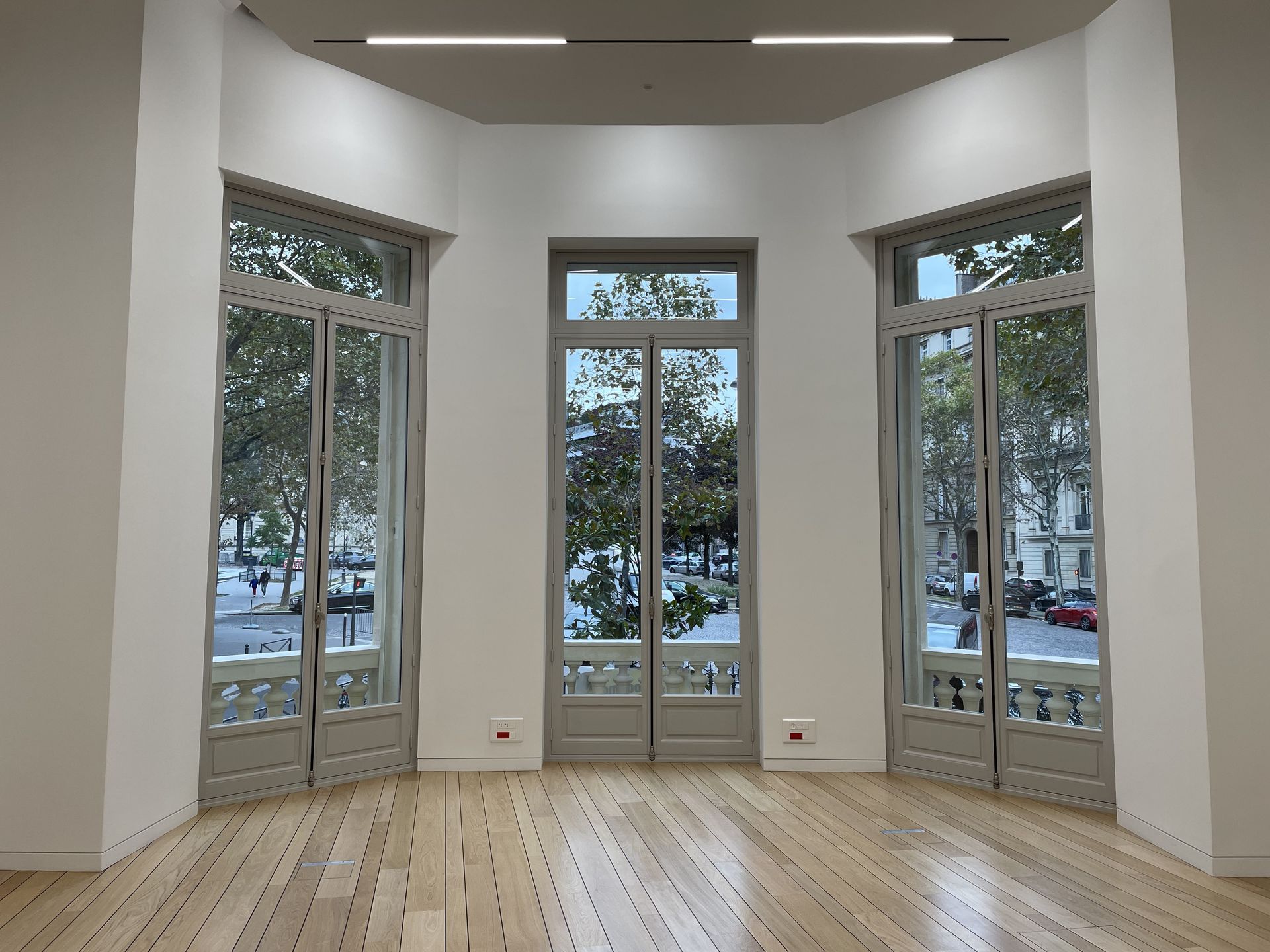 Trois grandes fenêtres cintrées aux cadres clairs illuminent une pièce aux sols en bois clair et aux murs blancs.
