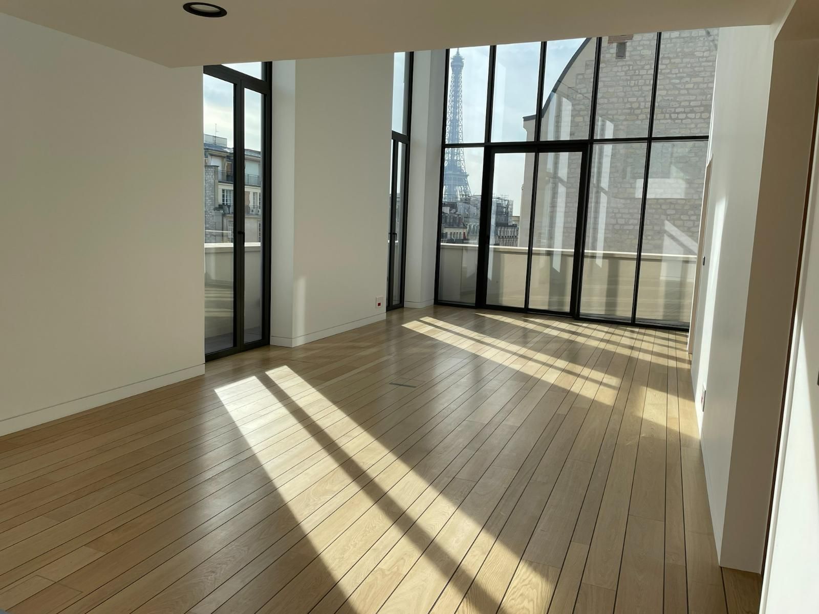 Pièce lumineuse et vide avec parquet, grandes fenêtres et balcon donnant sur la ville.